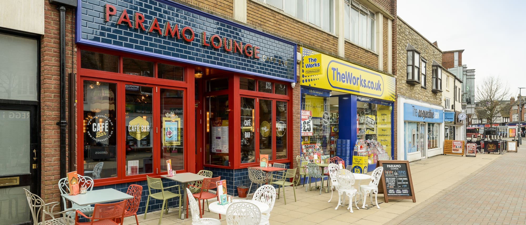 Street view of a café with outdoor tables and chairs beside the vibrant Paramo stationery shop, surrounded by other retail stores along a pedestrian walkway on a cloudy day.