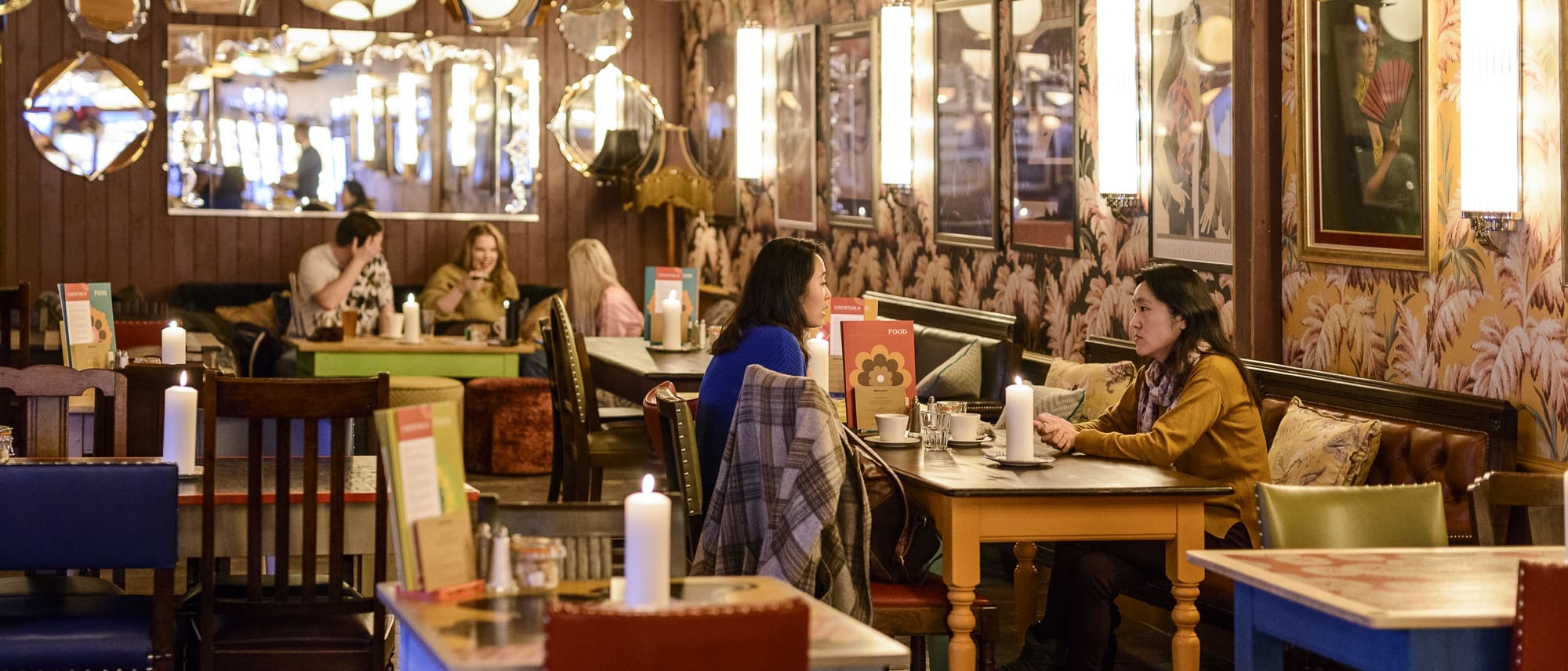 A cozy Paramo restaurant with warm lighting, colorful tables, and patterned wallpaper. Several groups of people sit at tables, talking and dining. Candles and menus decorate the tables, while mirrors and artwork hang on the walls.