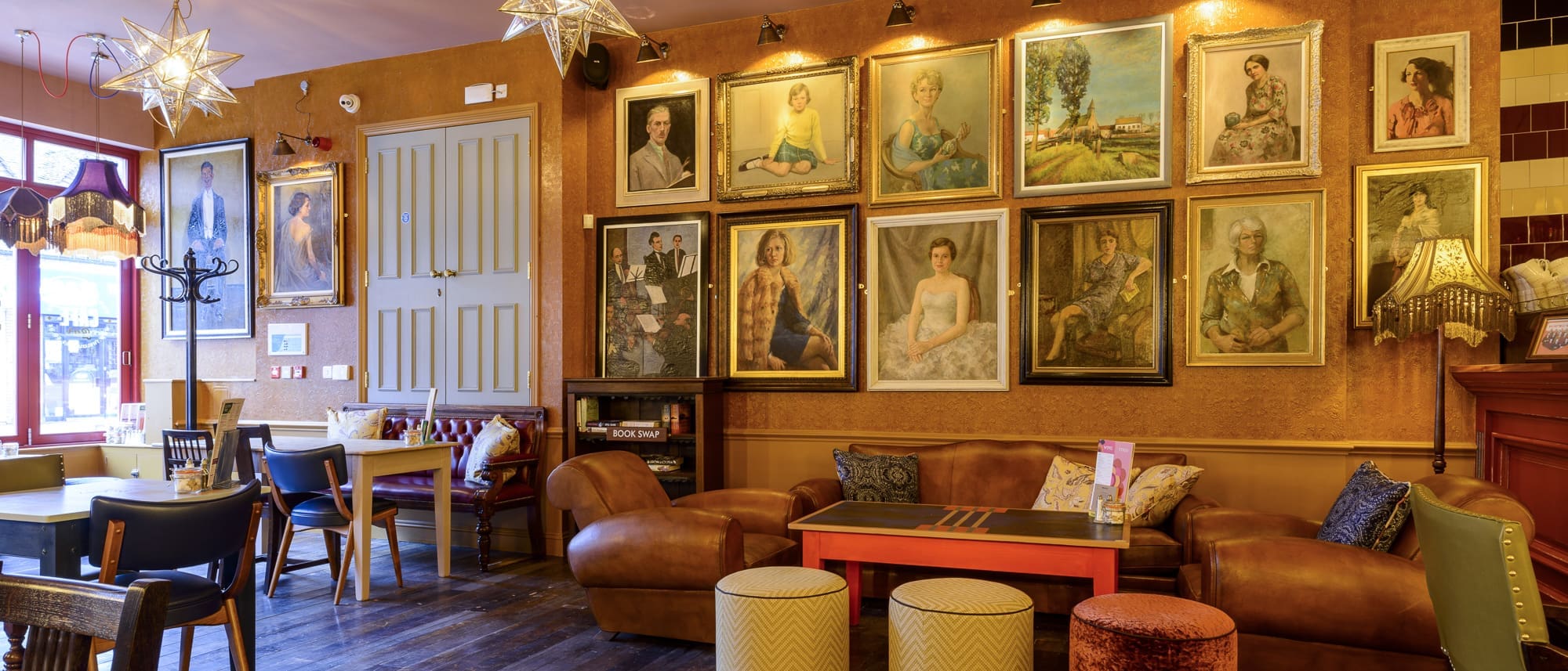 A cozy, warmly lit cafe with leather sofas, small tables, and mismatched chairs. The walls are decorated with many framed portraits and artwork inspired by Paramo landscapes, with star-shaped lights hanging from the ceiling.