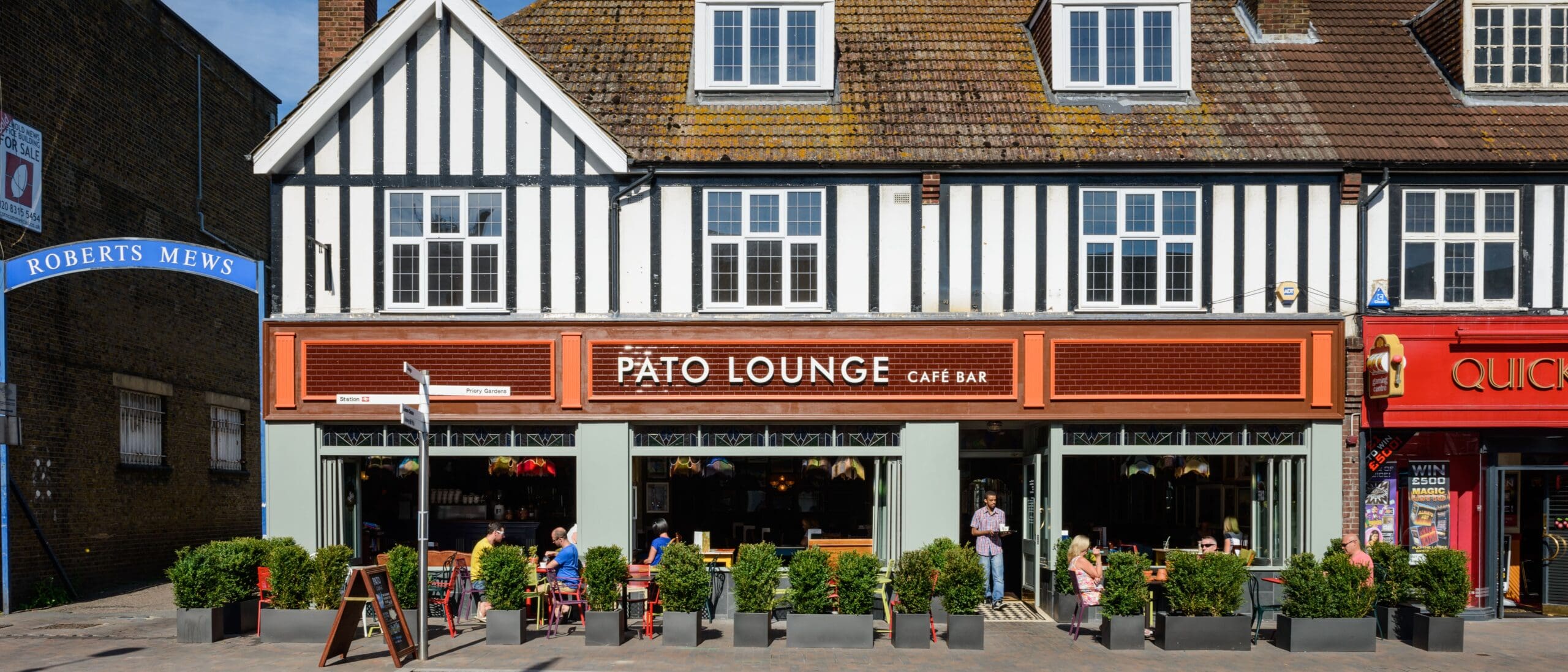 A street view of Pato Lounge Café Bar with outdoor seating, people dining, and a Tudor-style building facade. The vibrant Pato sign stands out, while a blue “Roberts Mews” sign is visible on the left and shops line either side.