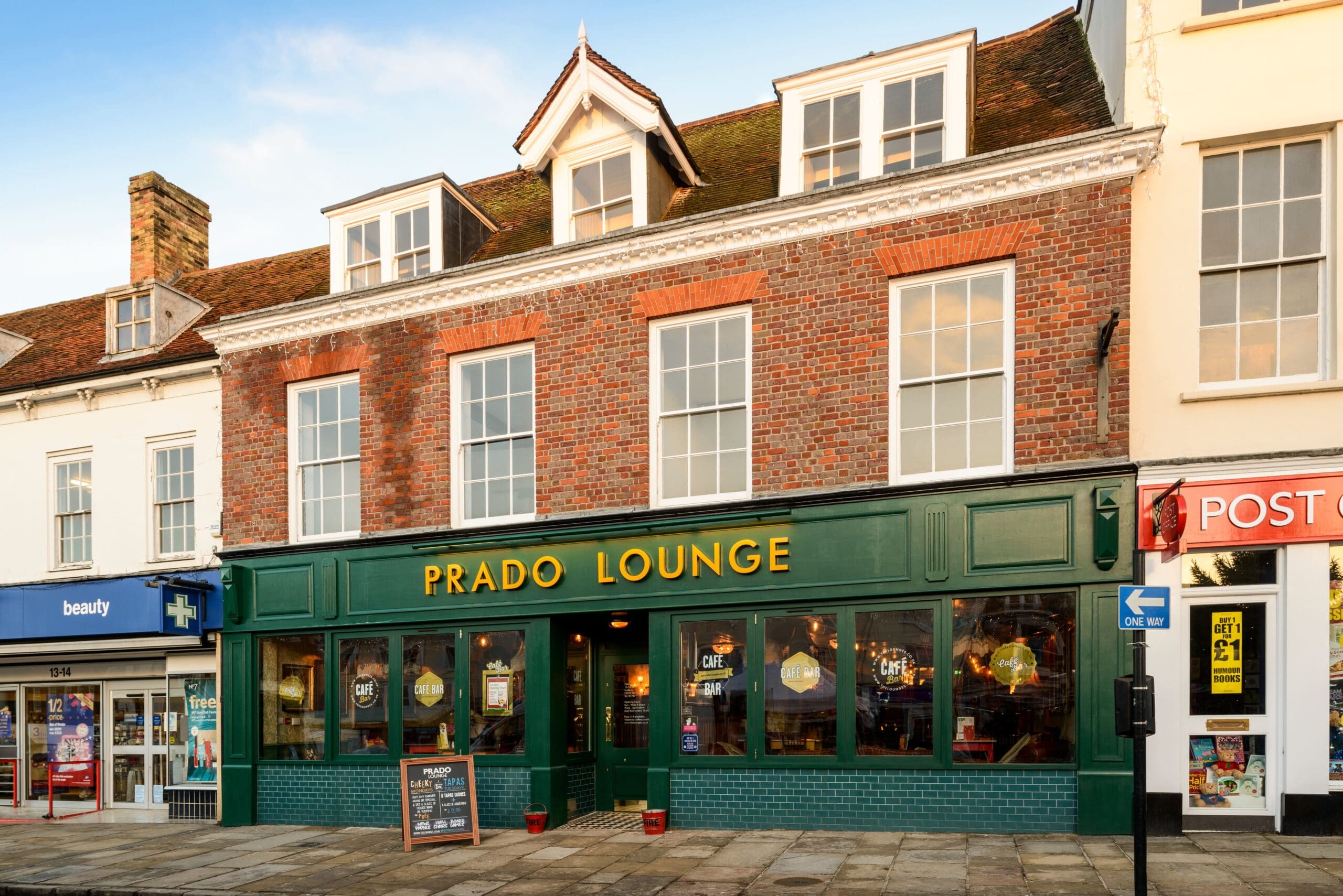 A brick building with large windows houses Prado Lounge, a lively Prado café-bar with a green and gold sign. Nestled between a beauty store and post office on a sunny day, it welcomes visitors with a sandwich board outside the entrance.