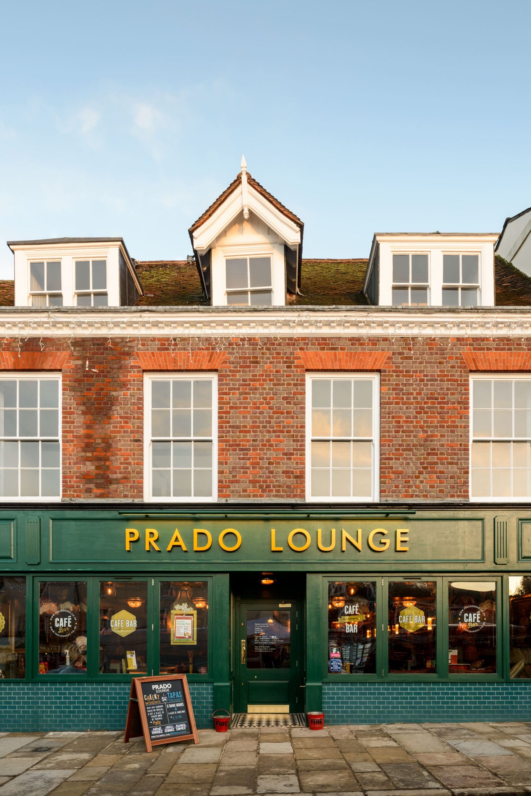 A brick building with large windows houses Prado Lounge, a café-bar. The green and yellow Prado storefront features bright signage, menu boards, and inviting, warmly-lit interiors visible through the windows.