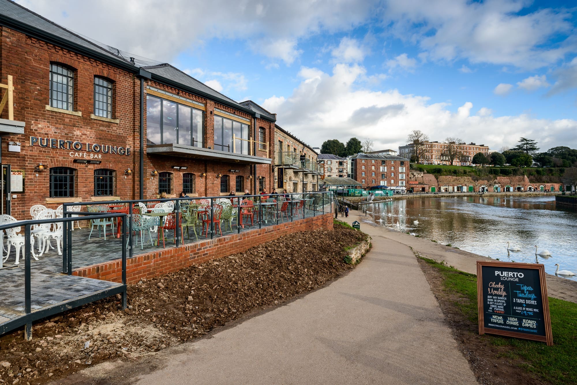 Puerto Lounge, a cozy brick café with outdoor seating, overlooks a riverfront path. Empty tables await guests on the terrace, beneath a partly cloudy sky and historic buildings—perfect for enjoying Puerto’s charm.