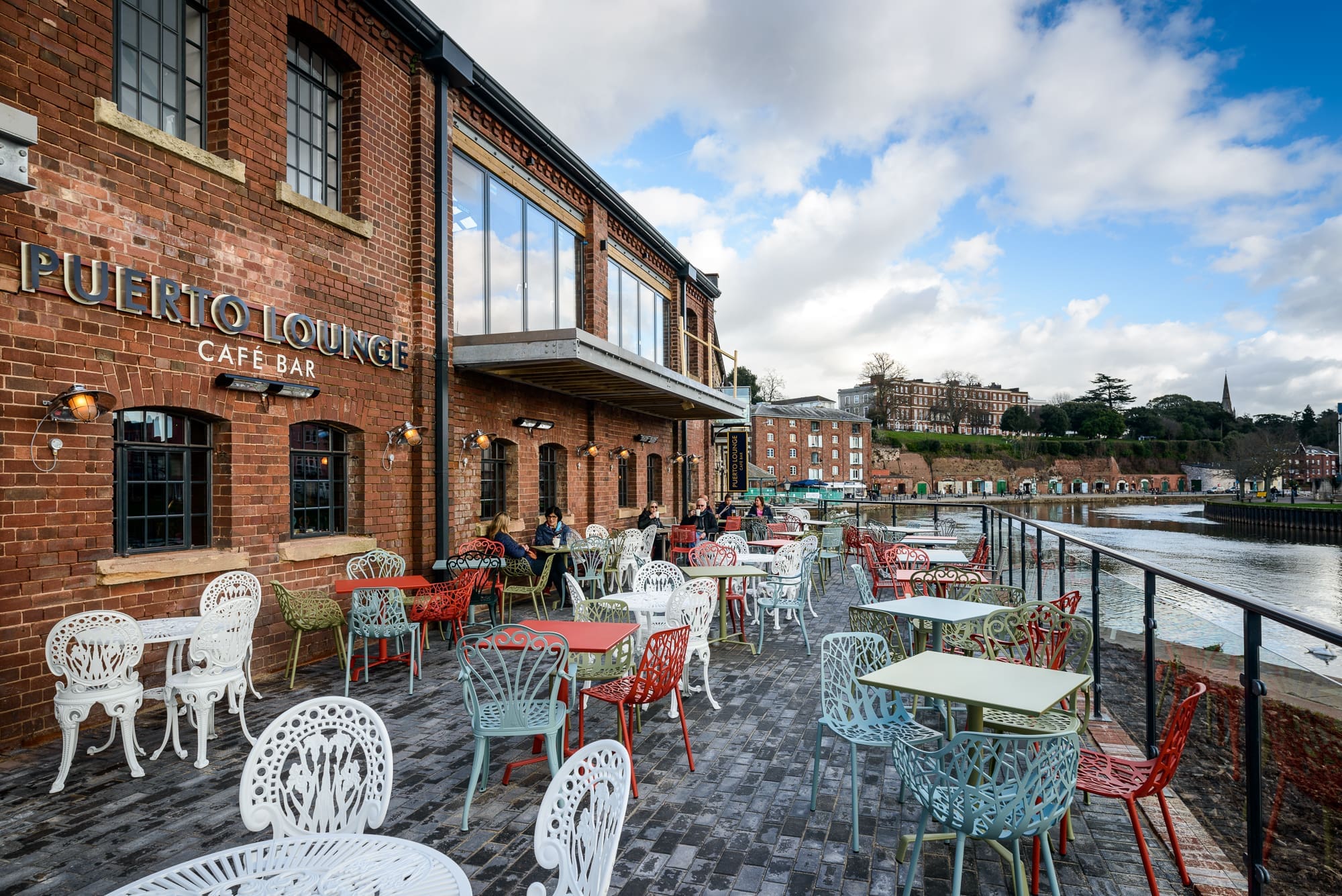 Relax at Puerto’s vibrant outdoor seating area, where colorful chairs and tables await on a brick patio beside the river, offering scenic views of neighboring historic buildings under a partly cloudy sky.