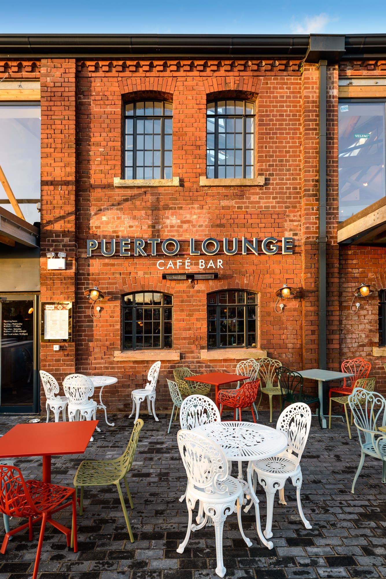 Brick building with large windows and a sign reading "Puerto Lounge Café Bar." Colorful metal tables and chairs are arranged outside on a cobblestone patio, creating a vibrant Puerto vibe under clear daylight.