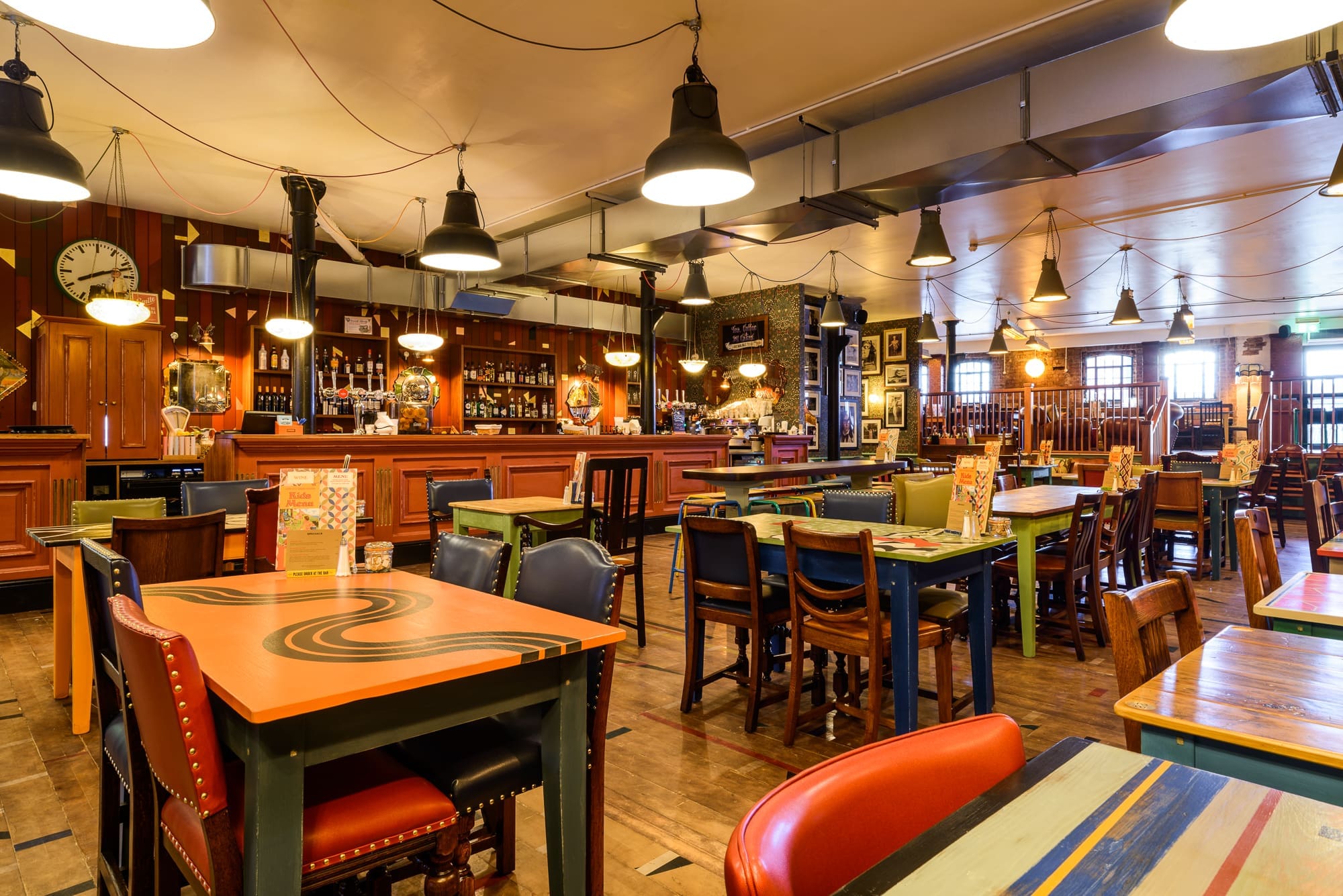 A brightly lit, eclectic pub interior in Puerto with colorful tables and mismatched chairs, a long wooden bar, various decorations on the walls, and menus placed on each table.