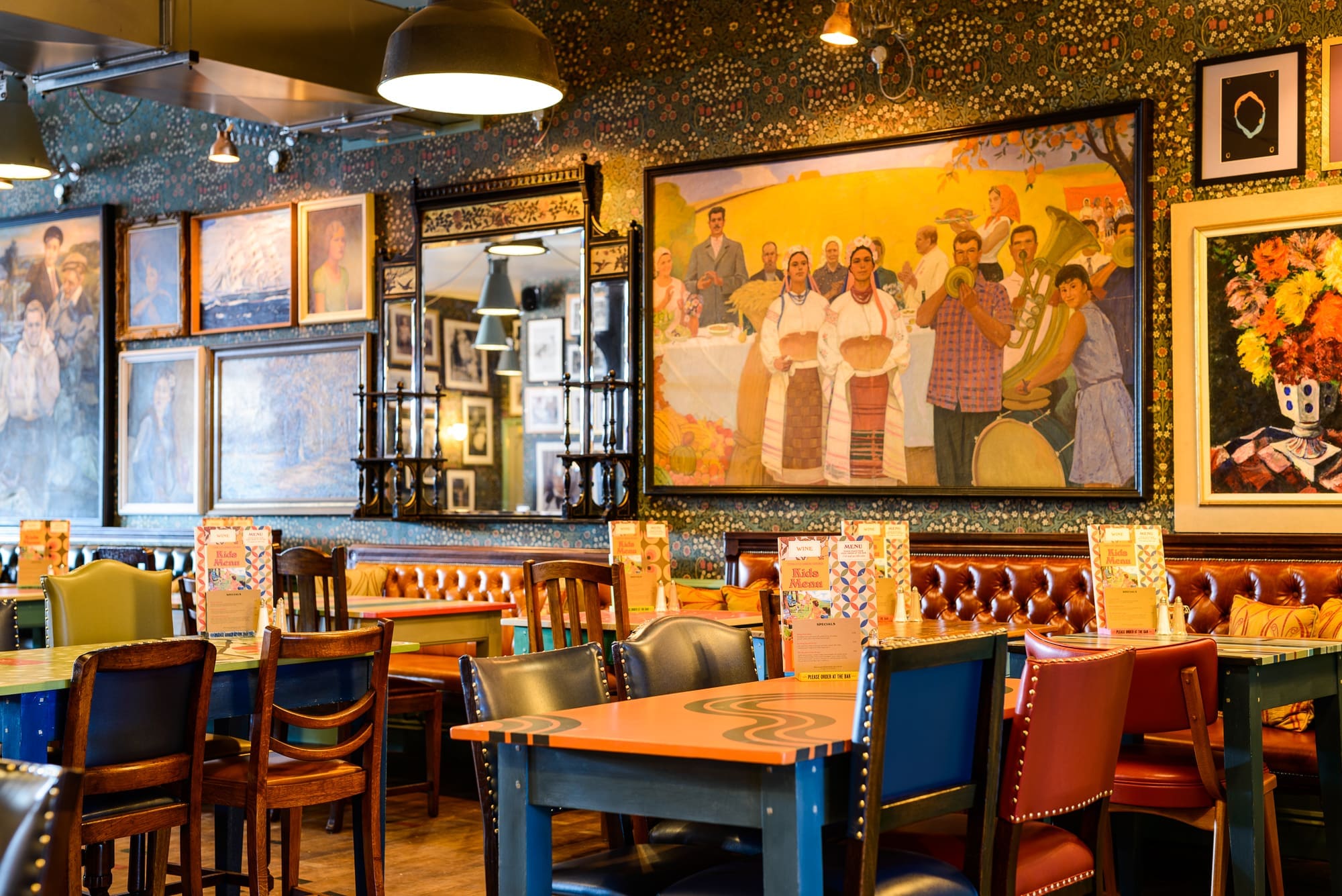 A colorful Puerto restaurant interior with eclectic chairs, wooden tables, and a wall decorated with large, vibrant paintings and framed artwork. Menus rest on each table; the space is well-lit and inviting.