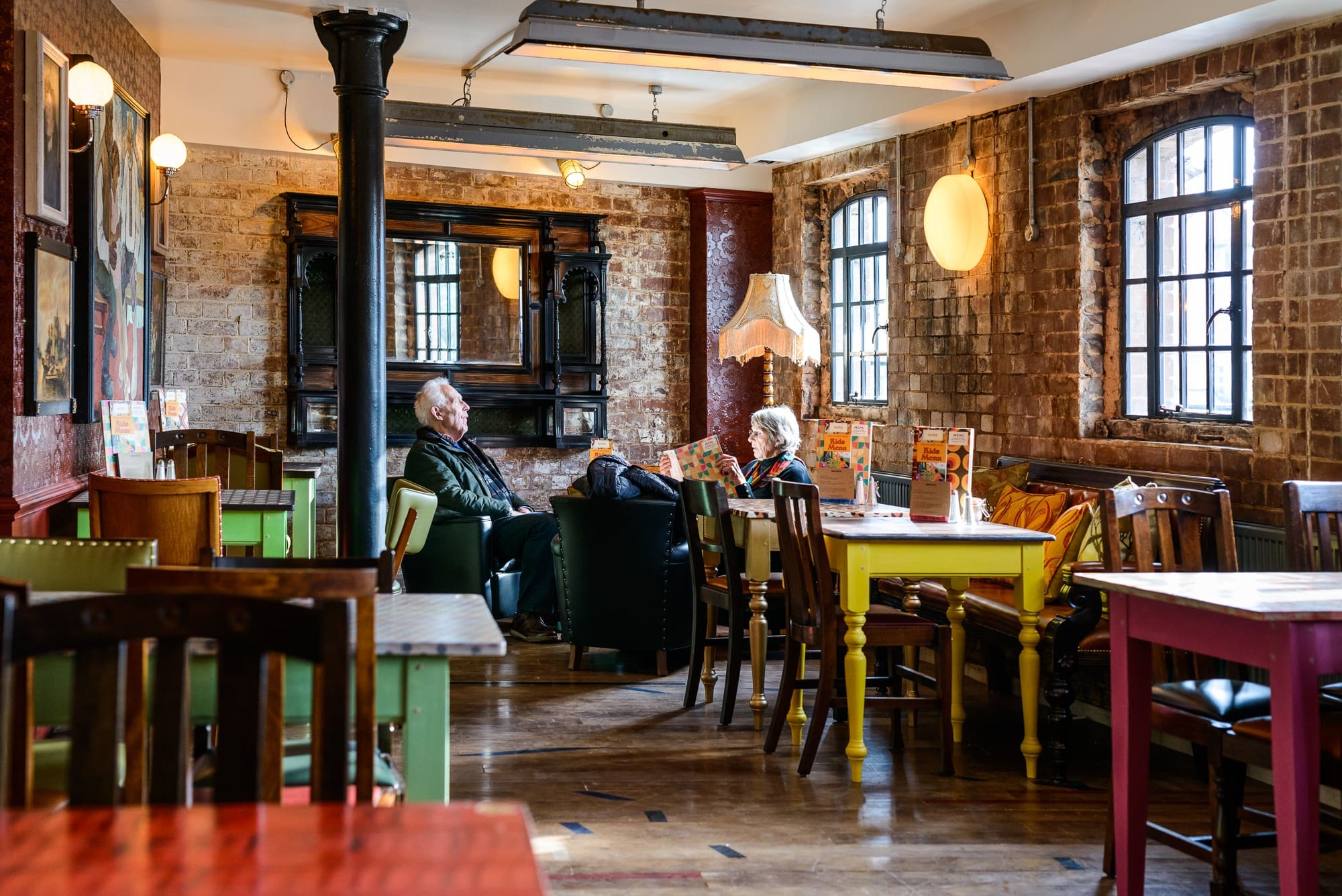 Two older adults sit and talk in a cozy, rustic café near the Puerto, with exposed brick walls, wooden floors, and mismatched furniture. Warm lighting and a relaxed atmosphere fill the space, featuring large windows and colorful tables.