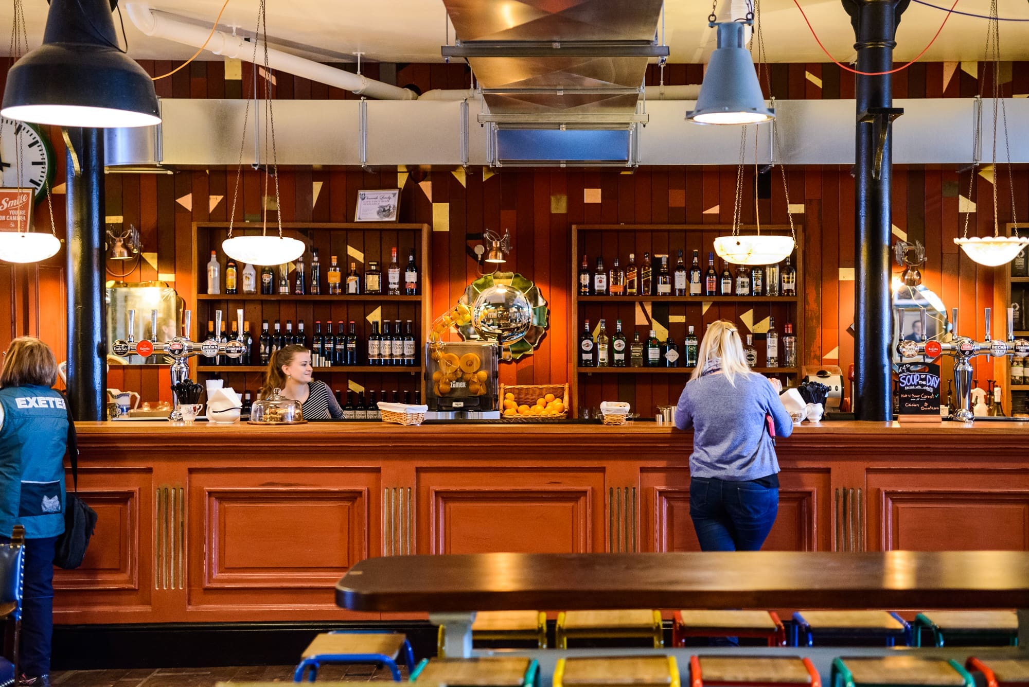 A brightly lit bar with red paneling, shelves of liquor bottles, two women at the counter, and rows of colorful stools in the foreground. Hanging lights and industrial decor evoke the vibrant spirit of a Puerto nightlife scene.
