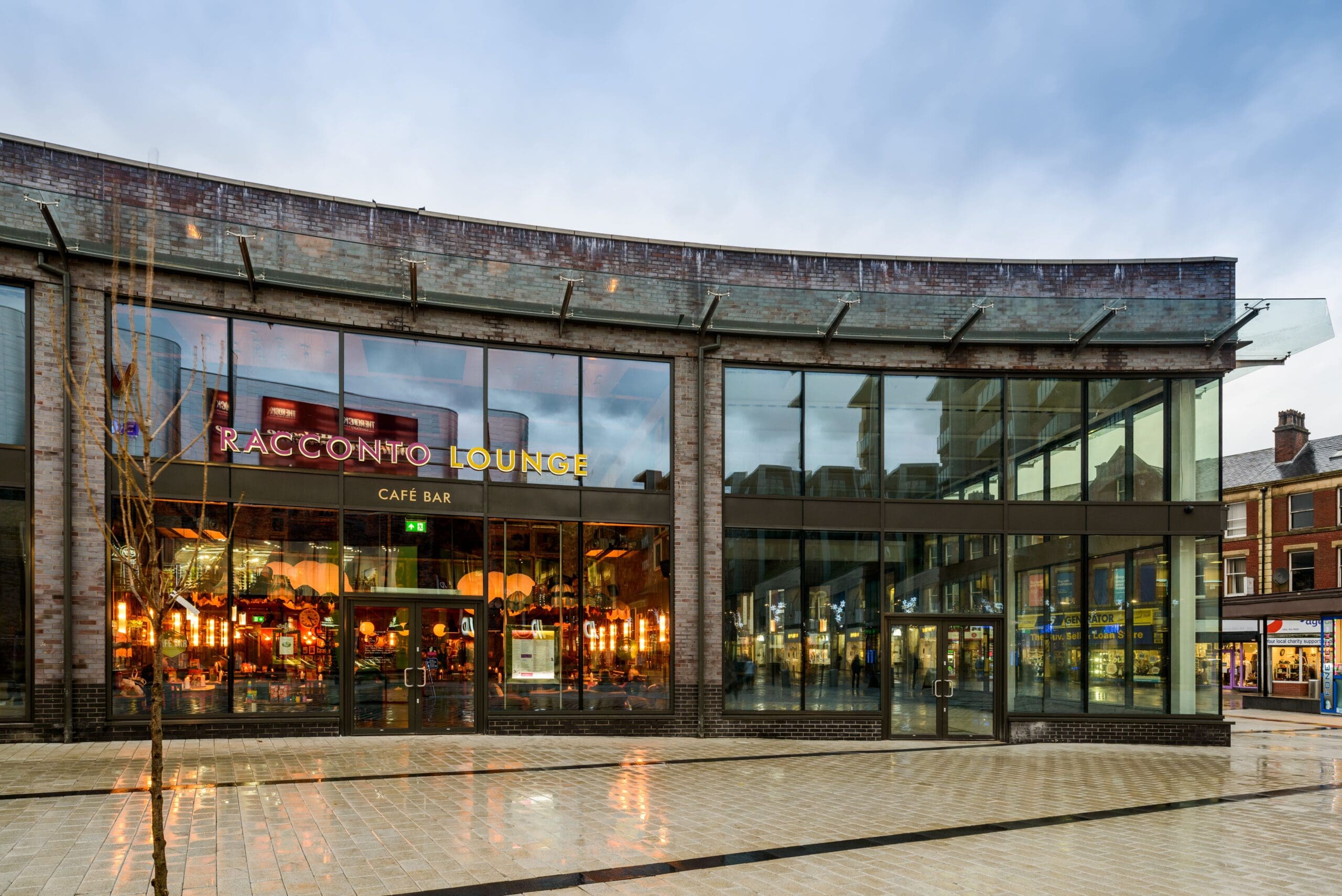 A modern glass-fronted building houses Racconto Lounge café bar, warmly lit inside. The wet pavement reflects the exterior on a cloudy day, with neighboring shops visible on the right.