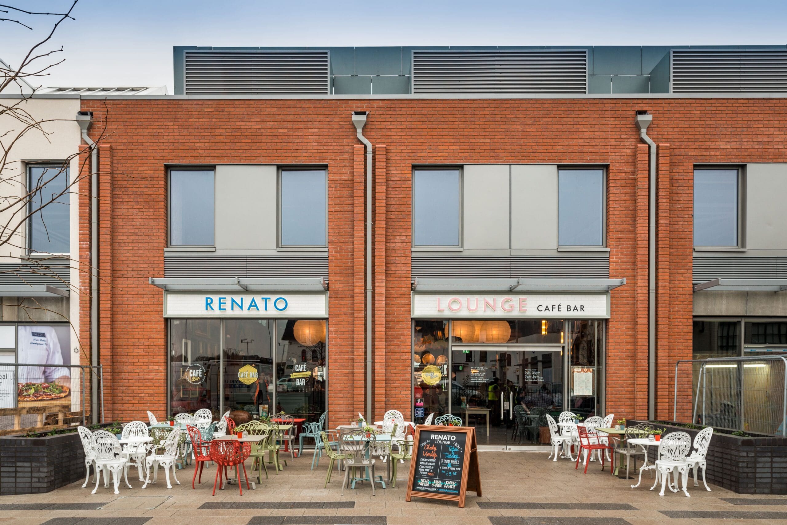 Outdoor seating area in front of Renato Lounge Café Bar, featuring colorful tables and chairs, a chalkboard menu, and large windows on a modern red-brick building. Experience the vibrant atmosphere at Renato's welcoming outdoor space.