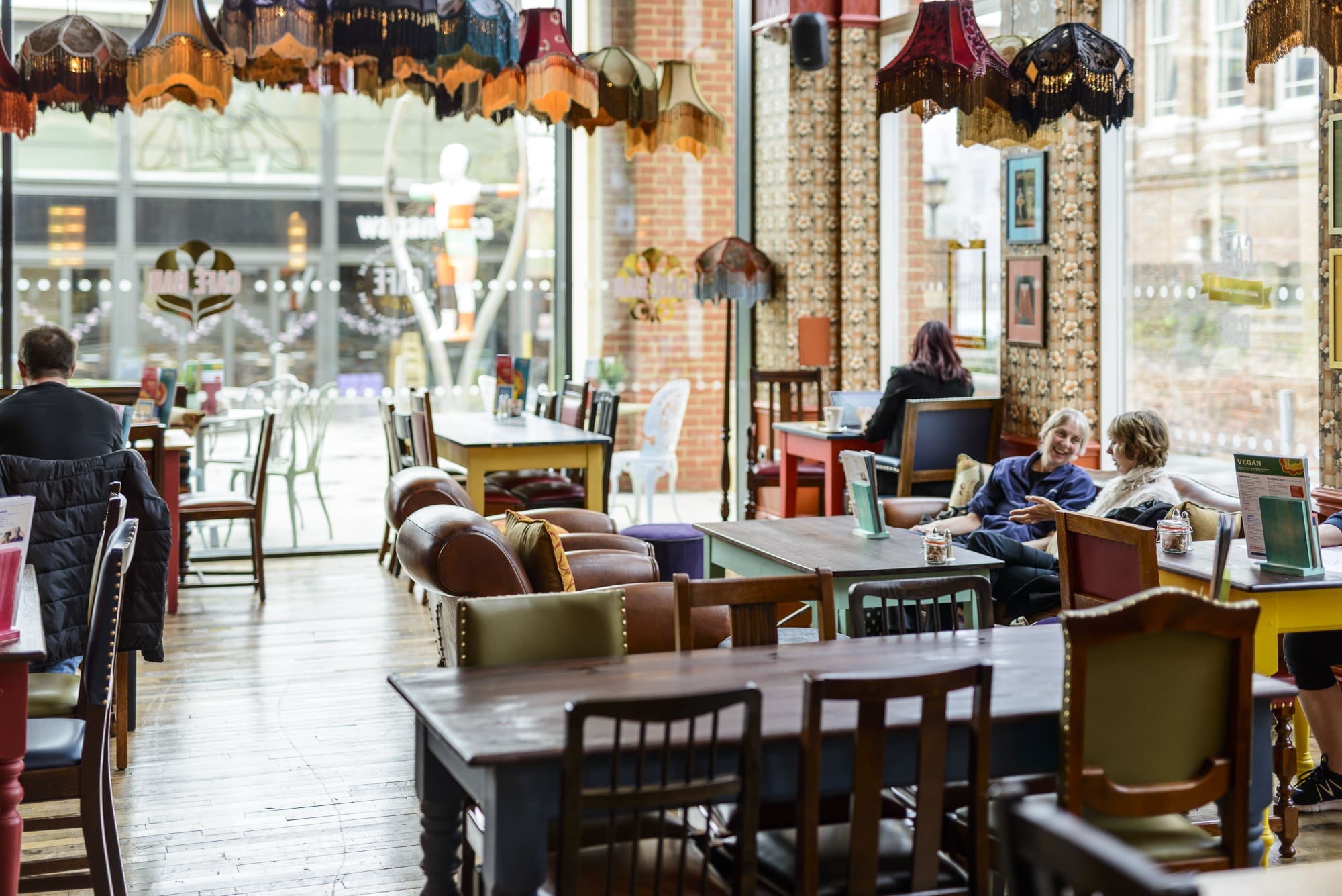 A cozy café with eclectic vintage decor, hints of Rococo flair, mismatched chairs, colorful lampshades, and large windows. People are sitting and chatting at tables, creating a warm, inviting atmosphere.