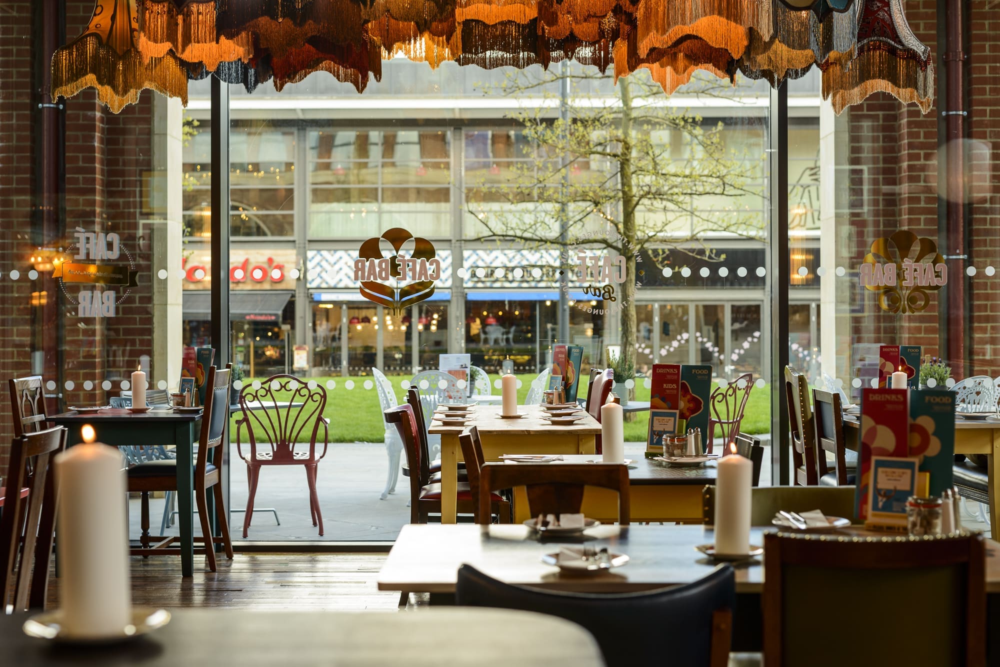 A cozy restaurant interior with Rococo-inspired colorful chairs and tables set for dining, lit by large white candles. A big window offers a view of a grassy area and shops outside. Daylight fills the space, creating a warm atmosphere.