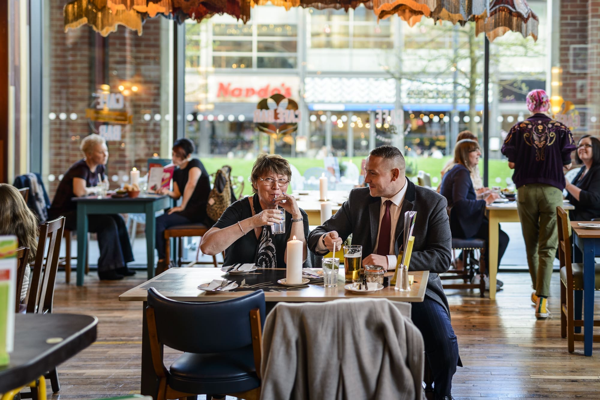 A man and woman sit at a restaurant table, enjoying a meal and drinks amid Rococo-inspired decor. Large windows reveal people and shops outside, while other diners in the background add to the lively atmosphere.