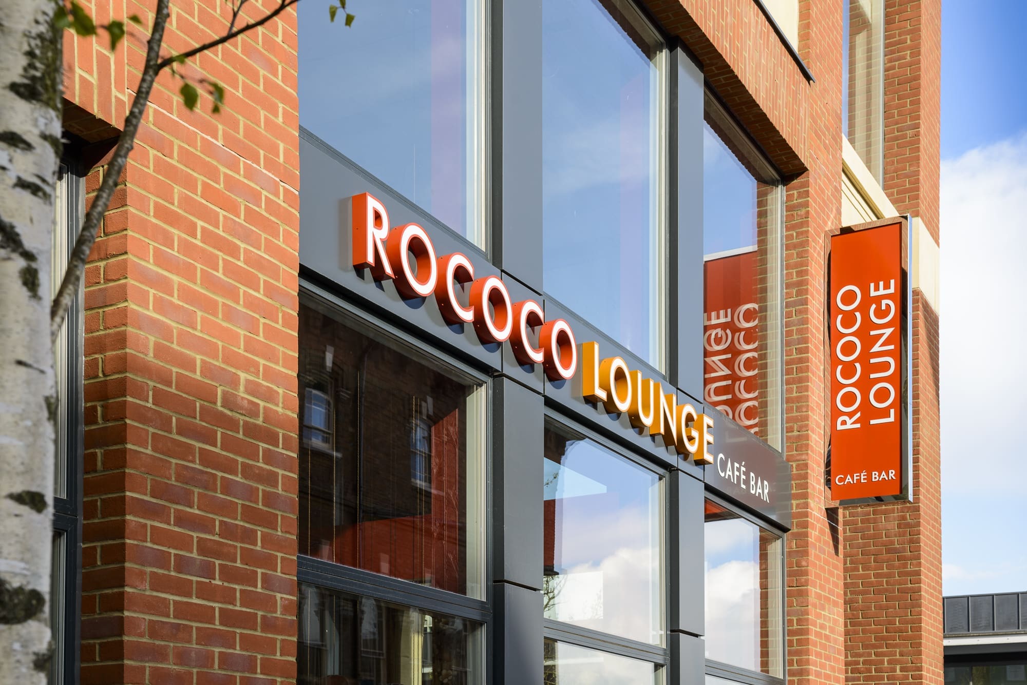 Exterior view of a brick building with large windows showcasing bright orange “ROCOCO LOUNGE CAFÉ BAR” signs. The stylish Rococo theme is echoed outside, with a tree trunk partially visible on the left side of the image.
