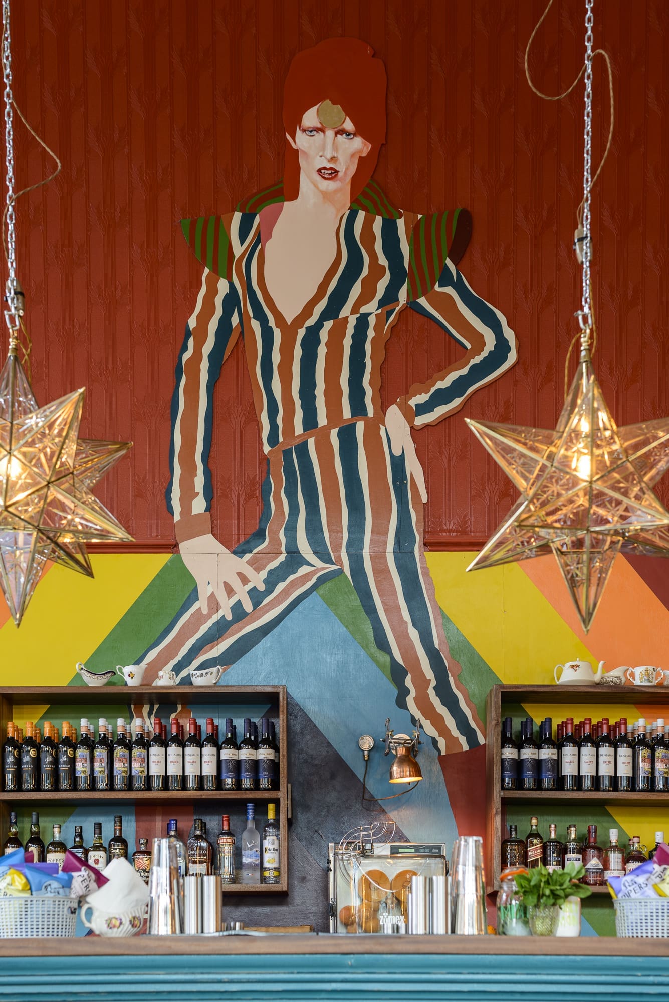 A colorful mural of a person in a striped suit, inspired by Rococo flair, is painted above a bar with shelves of assorted bottles. Two star-shaped pendant lights hang in the foreground.