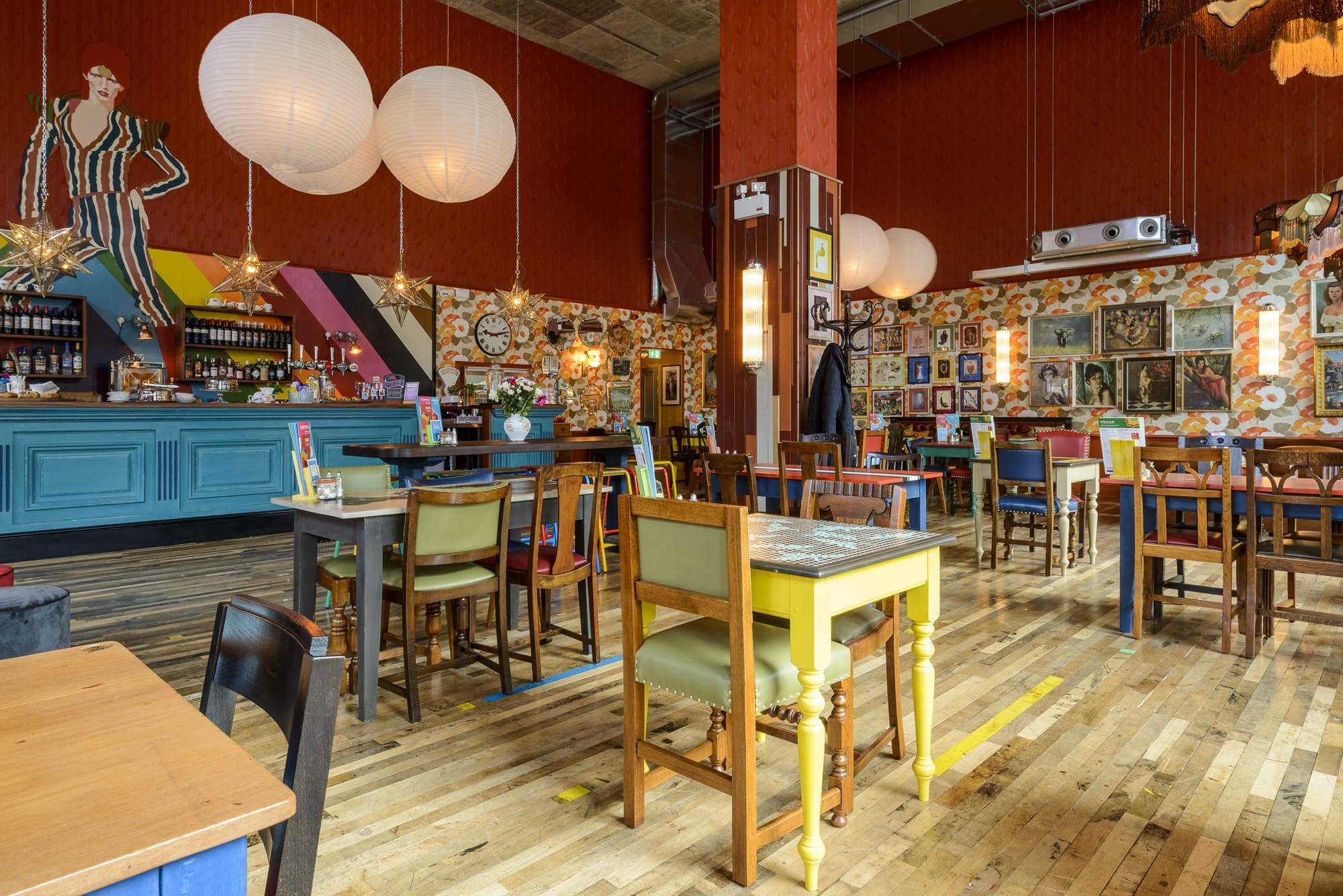 A colorful, eclectic cafe interior with mismatched chairs and tables, bright patterned walls, Rococo-inspired accents, hanging globe lights, and various framed art pieces and decorations throughout the space.
