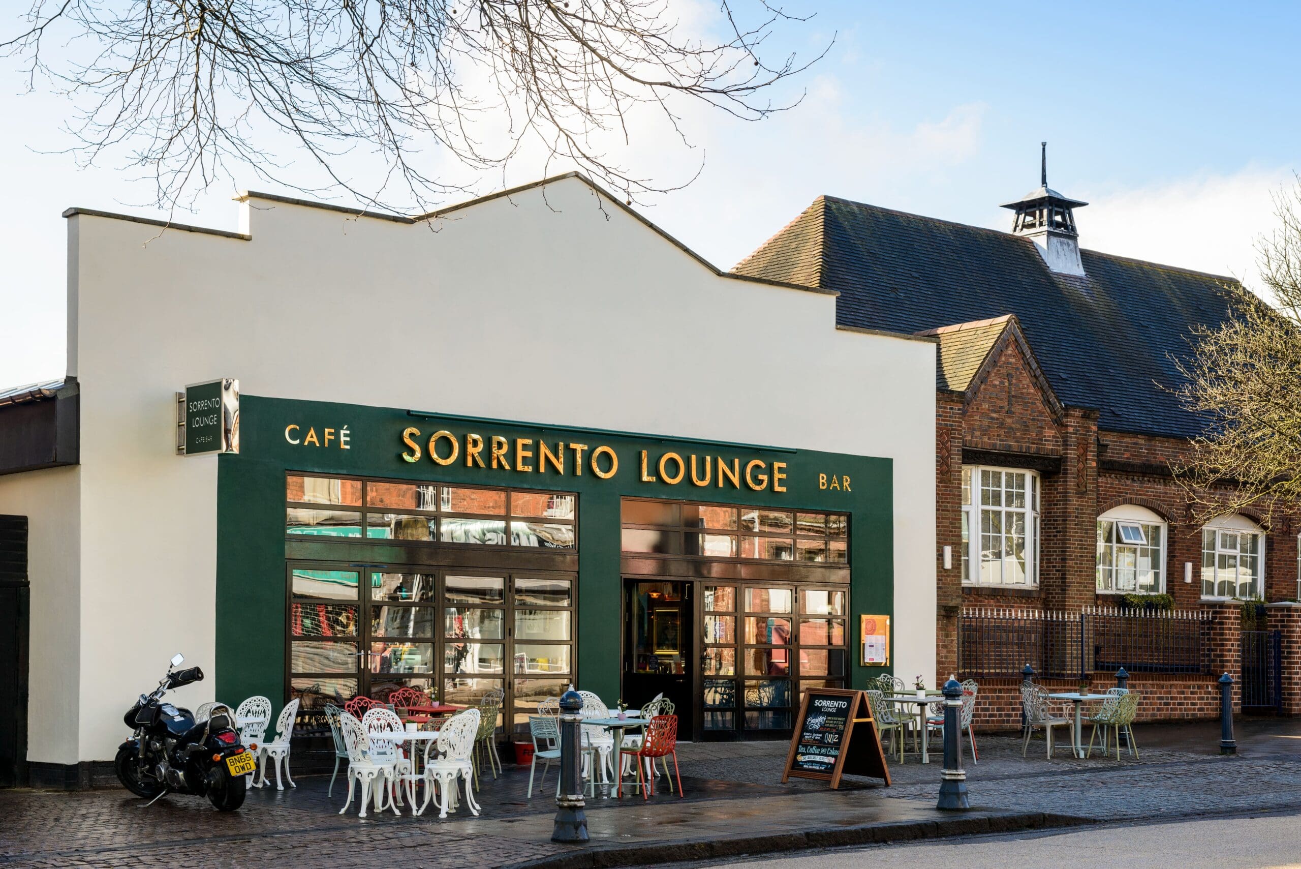 Sorrento Lounge is a stylish café-bar with green trim, outdoor seating, and a chalkboard menu. Its distinctive roofline and large windows stand out, while a motorcycle parked nearby adds to the laid-back Sorrento vibe on a sunny day.