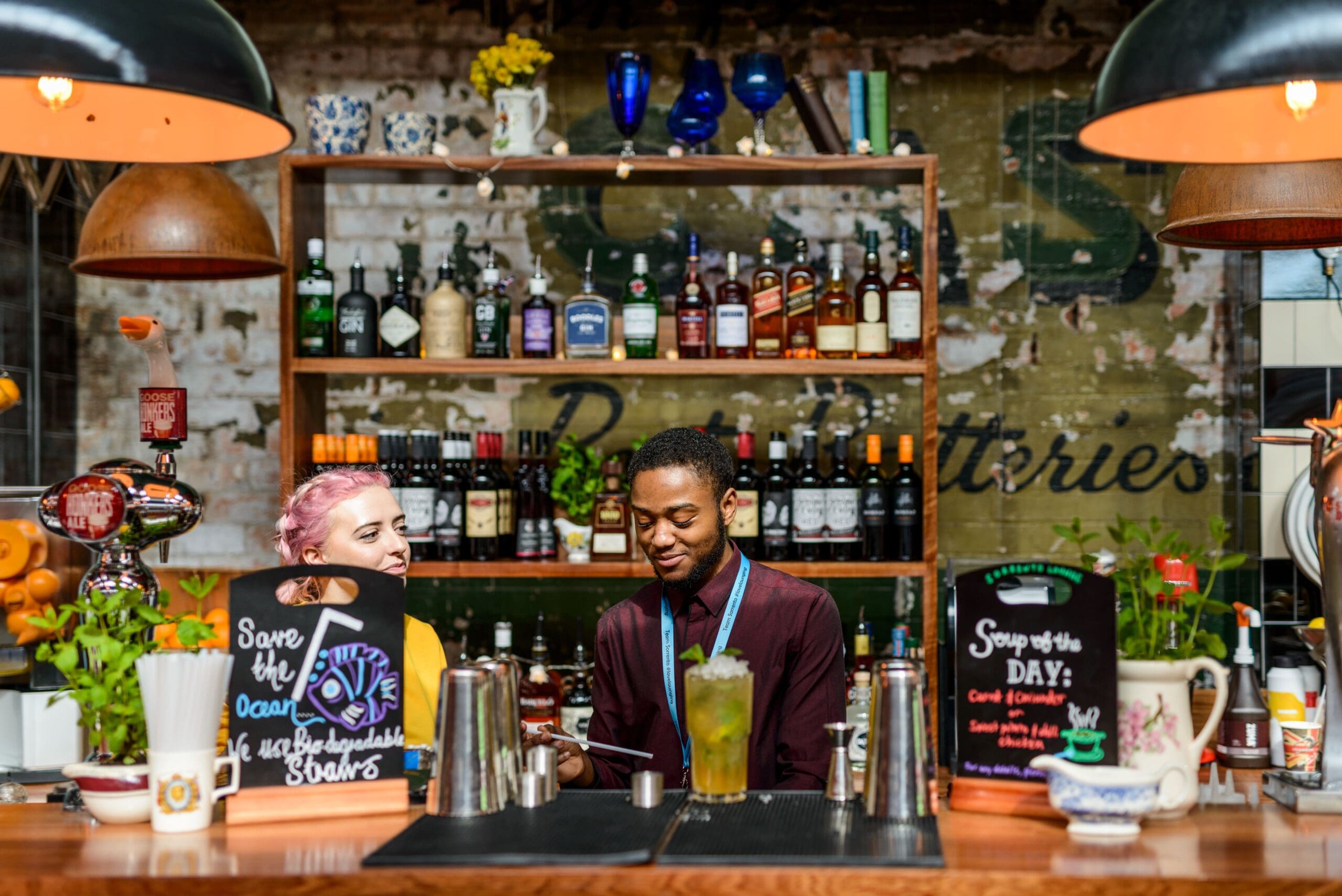 Two bartenders, one with pink hair and one with short dark hair, smile and chat behind a colorful Sorrento-inspired bar filled with bottles, plants, drink menus, and mixers. The atmosphere looks vibrant and inviting.