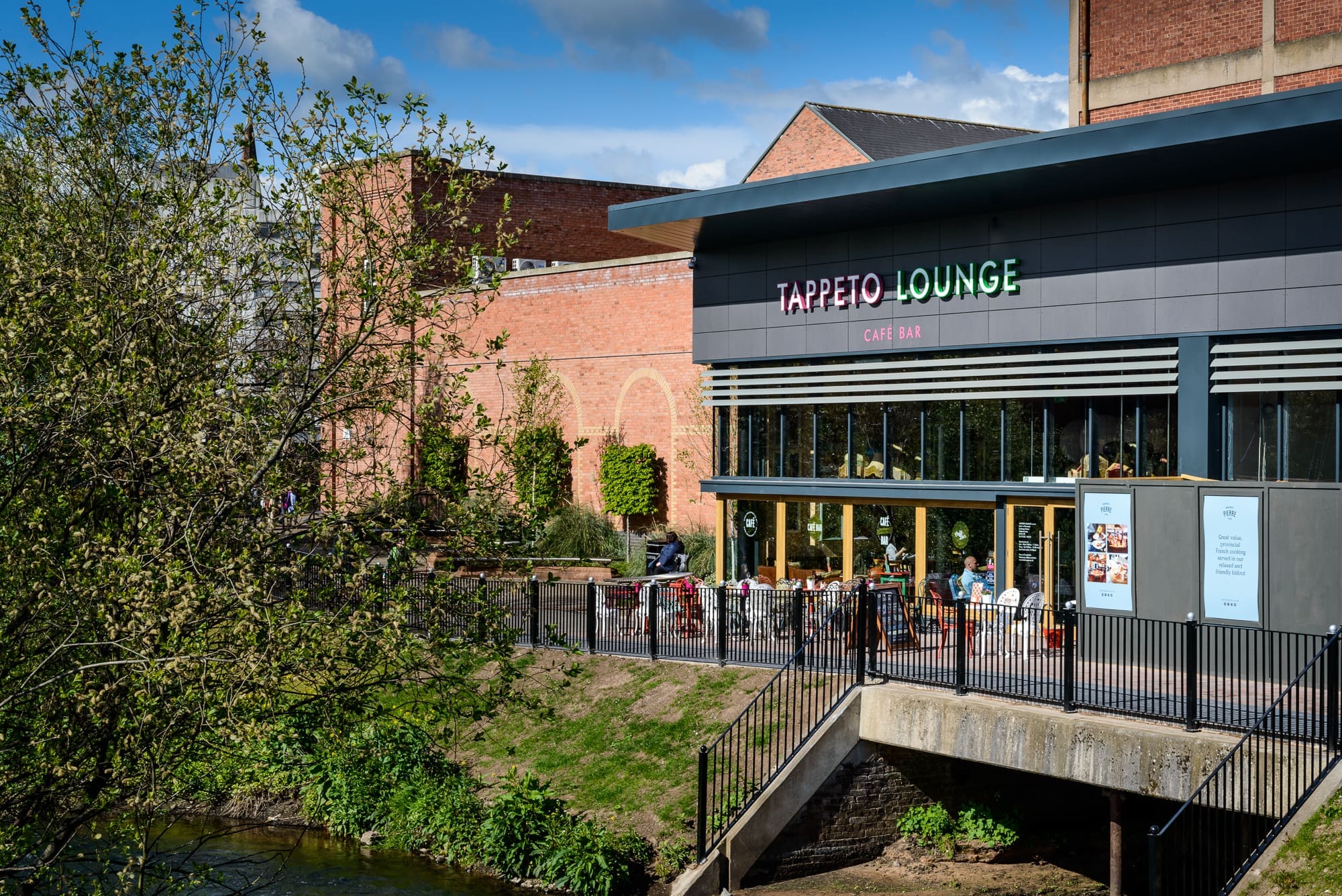 Tappeto Lounge is a modern café bar with outdoor seating overlooking the river. Guests enjoy dining under the sun, surrounded by red brick buildings and greenery, with large windows enhancing Tappeto's inviting atmosphere.