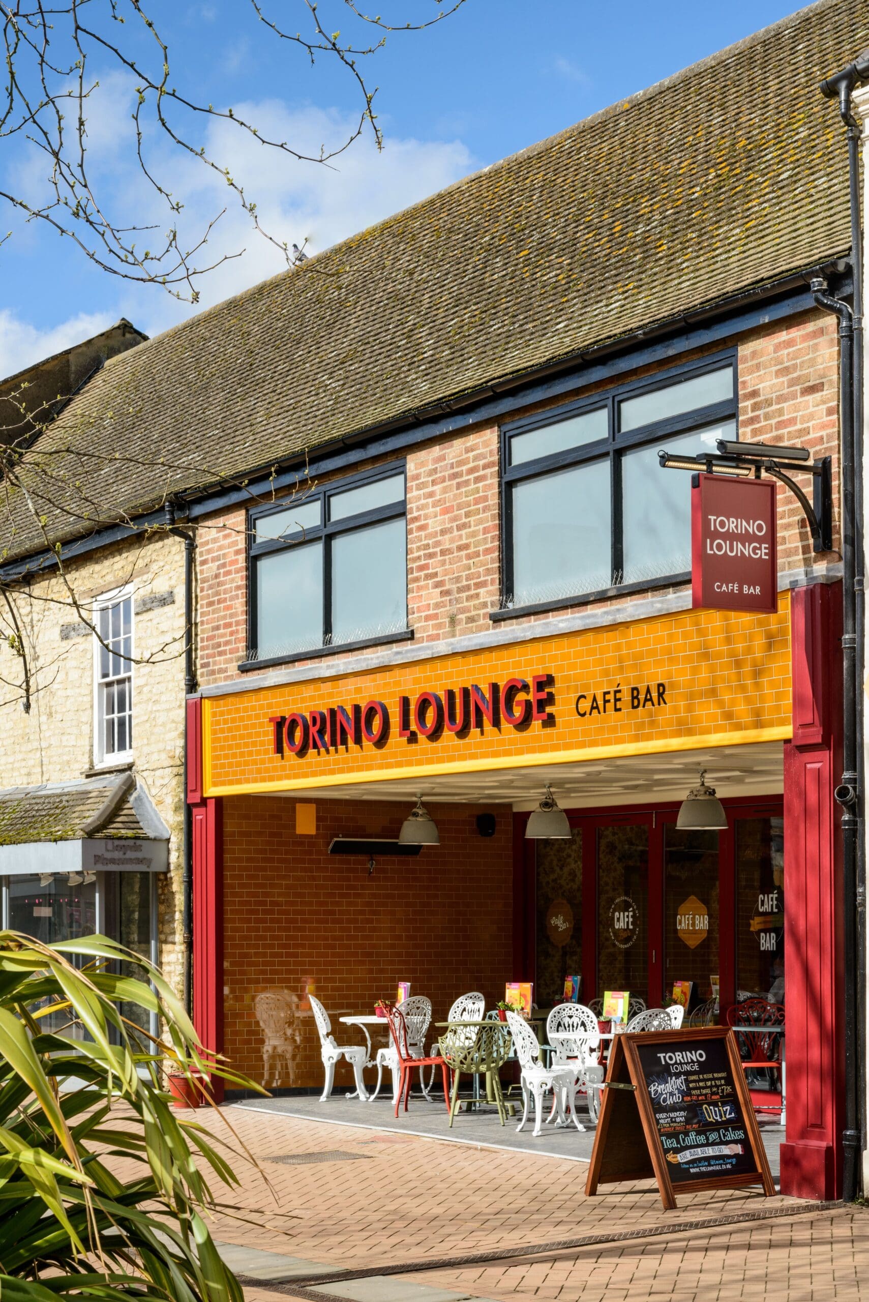 Torino Lounge is a café bar with outdoor seating and vibrant orange and yellow signage. Its Torino-inspired brick exterior gleams on a sunny day, with tables, chairs, and a chalkboard menu welcoming guests at the entrance.