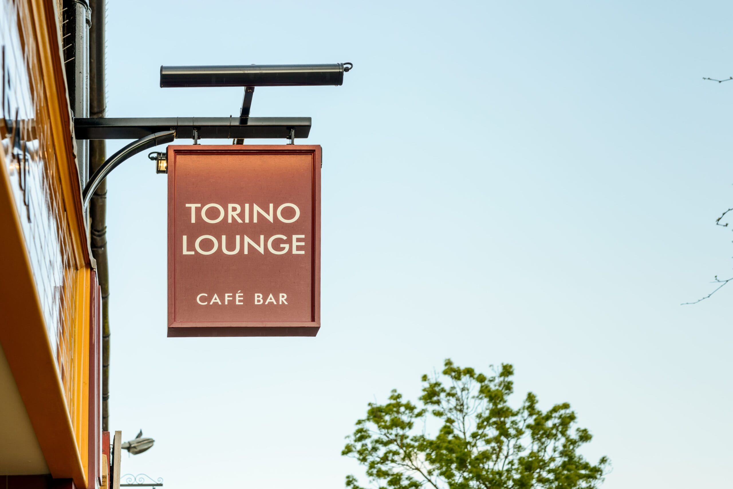 A red "Torino Lounge Café Bar" sign hangs from a building against a clear sky, with part of a tree in the background, capturing the inviting charm of Torino.
