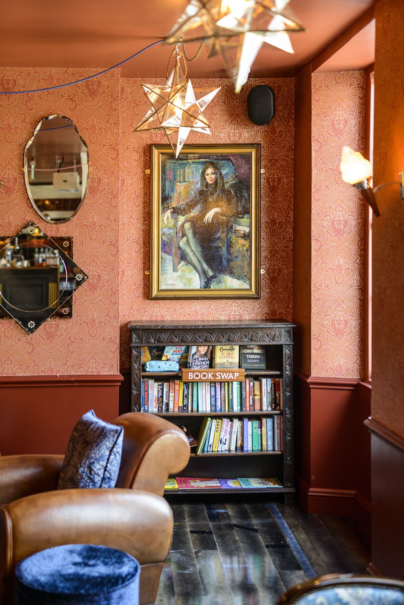 A cozy reading nook with patterned orange walls, a bookshelf filled with books labeled "Book Swap," a brown leather armchair, a framed portrait of a woman, and star-shaped ceiling lights.