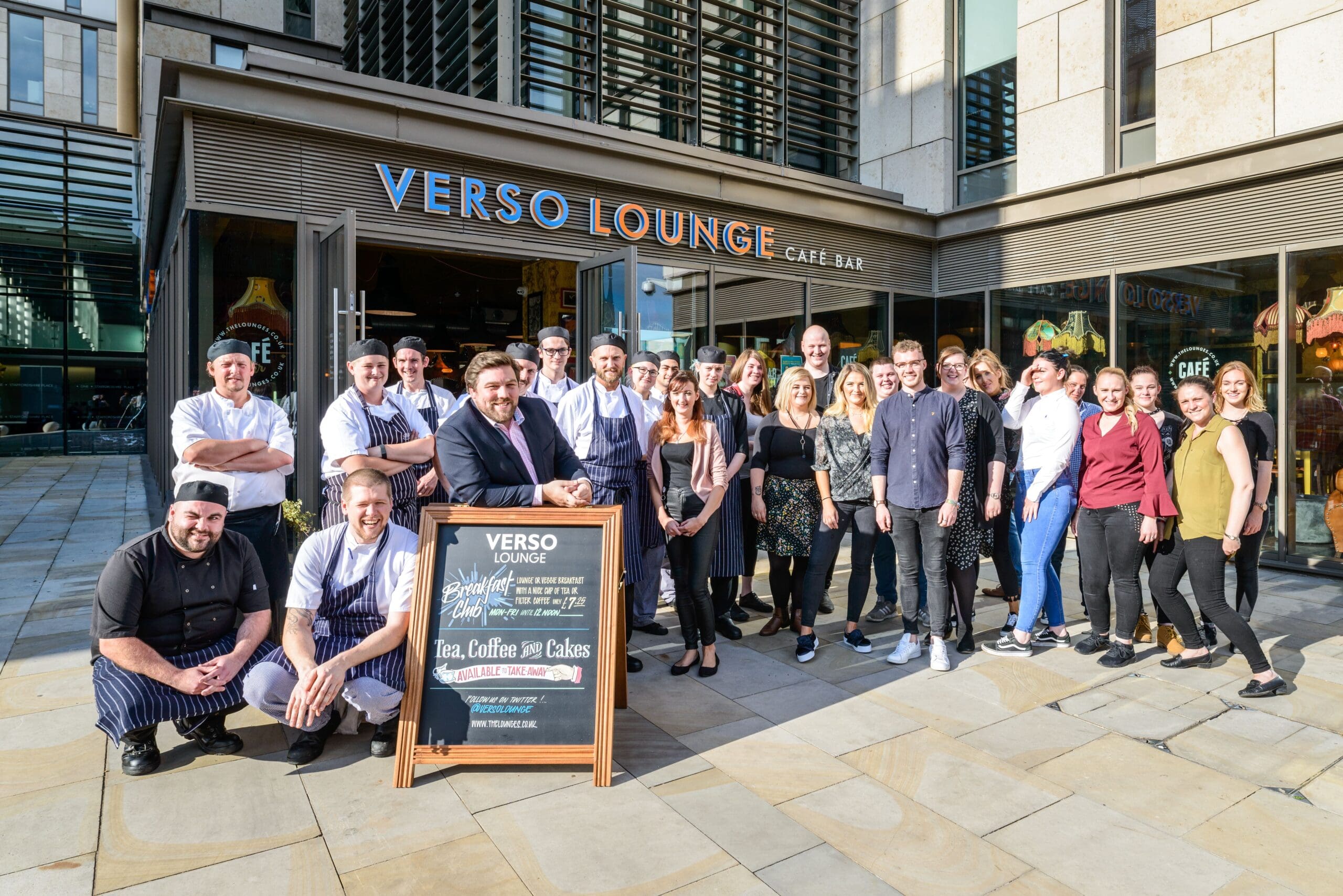 A large group of people, some in Verso chef uniforms and others in casual or work attire, stand smiling outside Verso Lounge café bar. A chalkboard sign with menu items is displayed in front of the group on a sunny day.