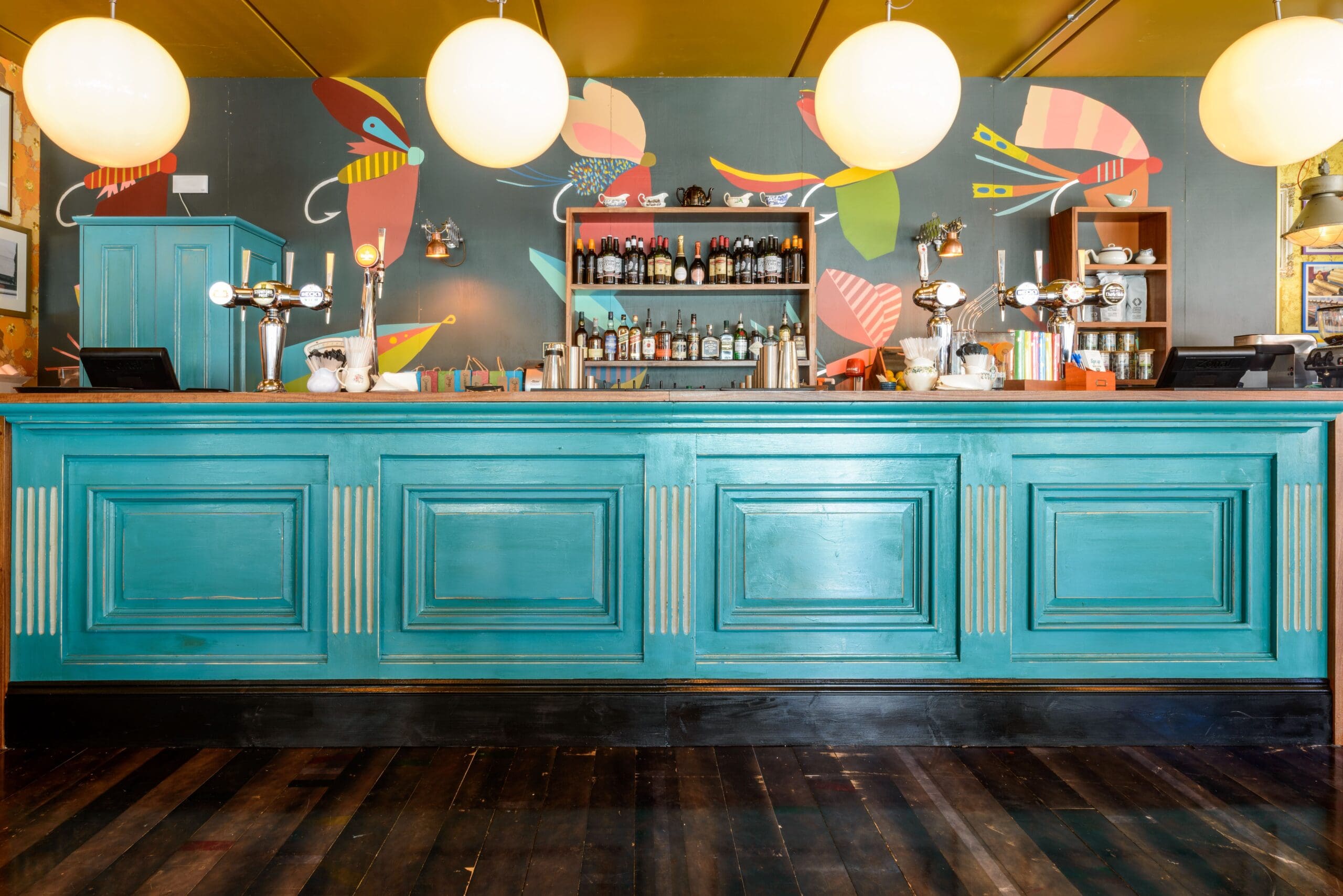 A vibrant bar with a turquoise counter from Verso, globe pendant lights overhead, and colorful abstract wall art behind shelves stocked with bottles and glassware on a wooden floor.