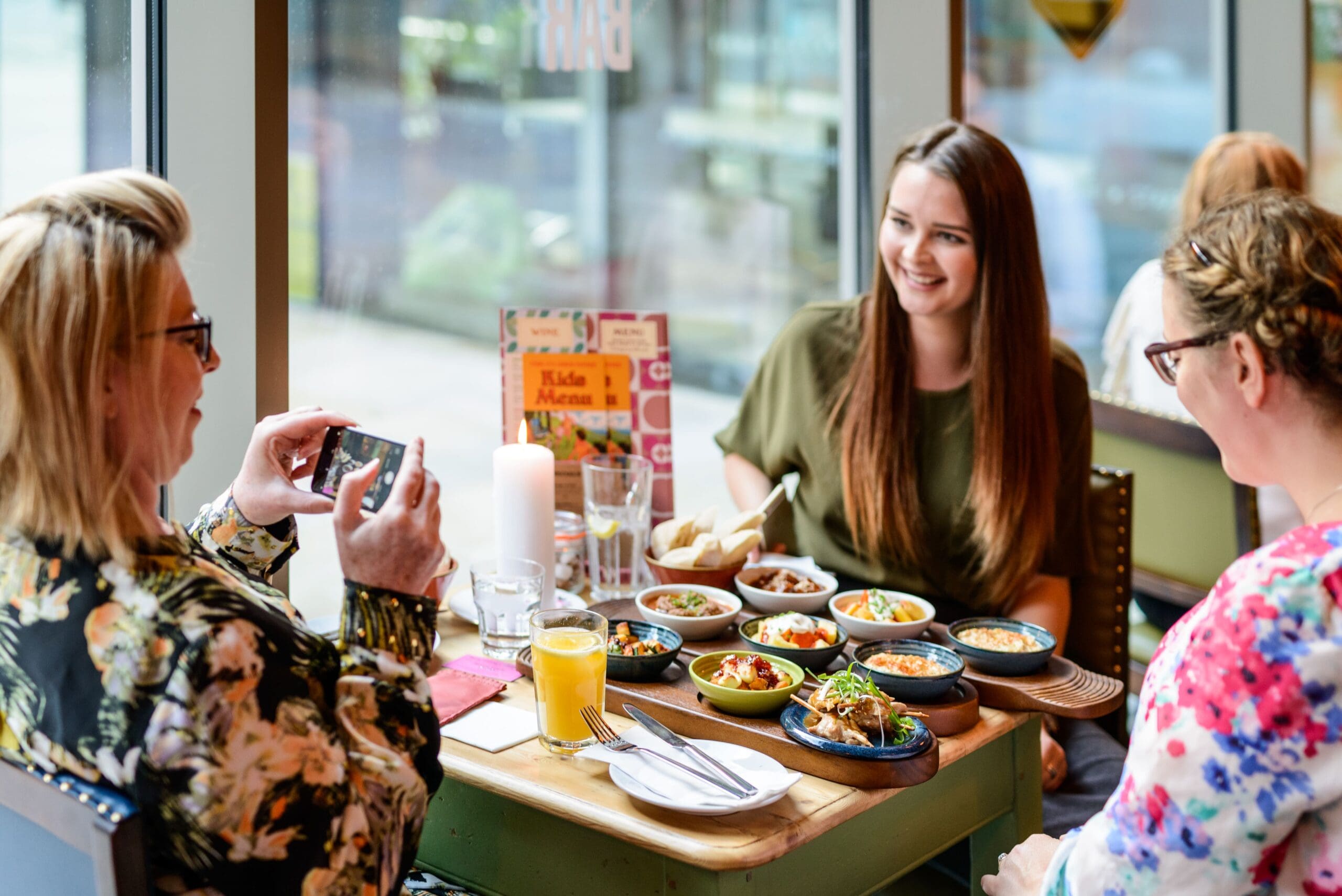 Three women sit at a Verso restaurant table with various small dishes and drinks, smiling and chatting as one woman snaps a photo of the food. Large windows fill the space with natural light in the background.