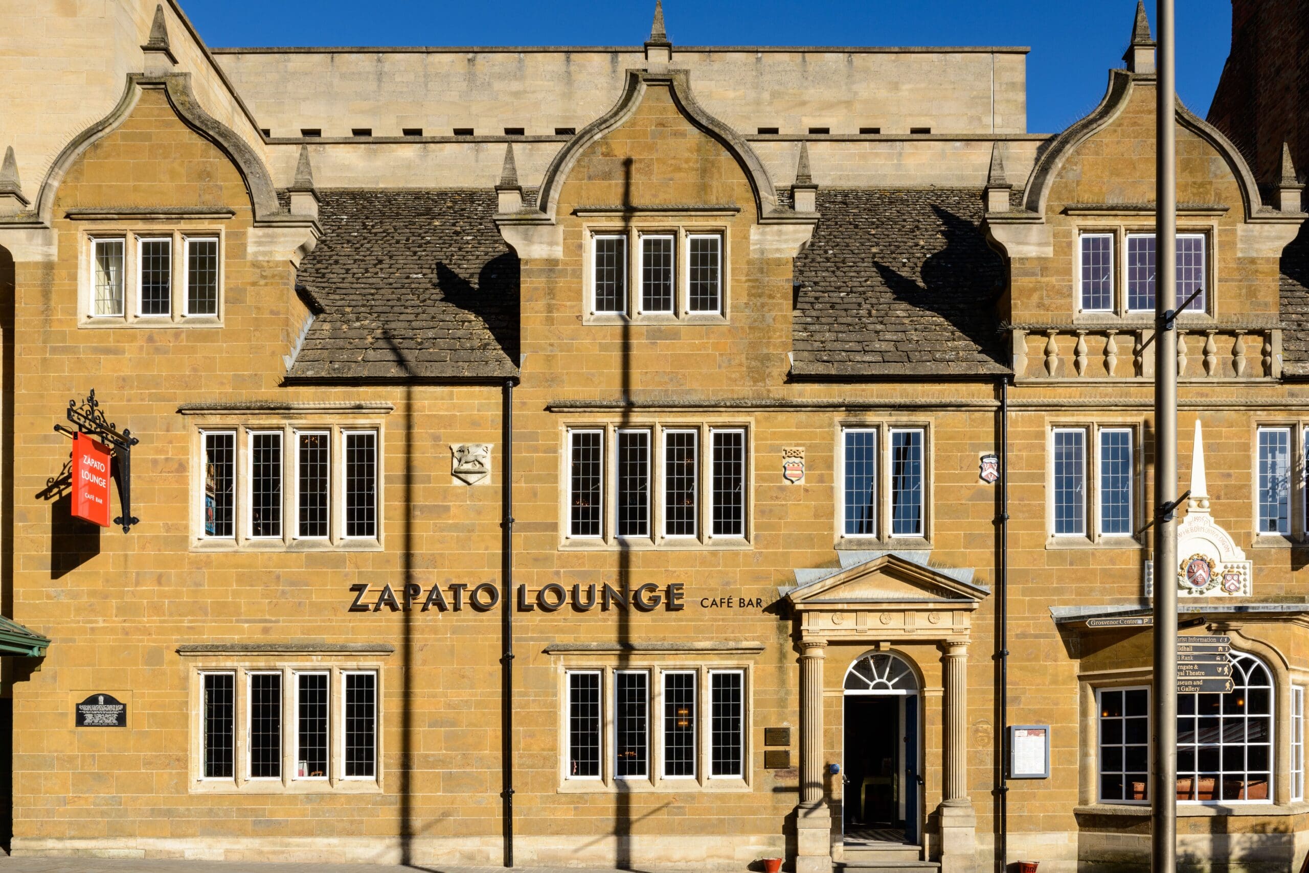 Zapato Lounge Café Bar, housed in a historic stone building with large white-framed windows and ornate gables, sits under a clear blue sky. A red Zapato sign and a streetlamp welcome guests at the entrance.