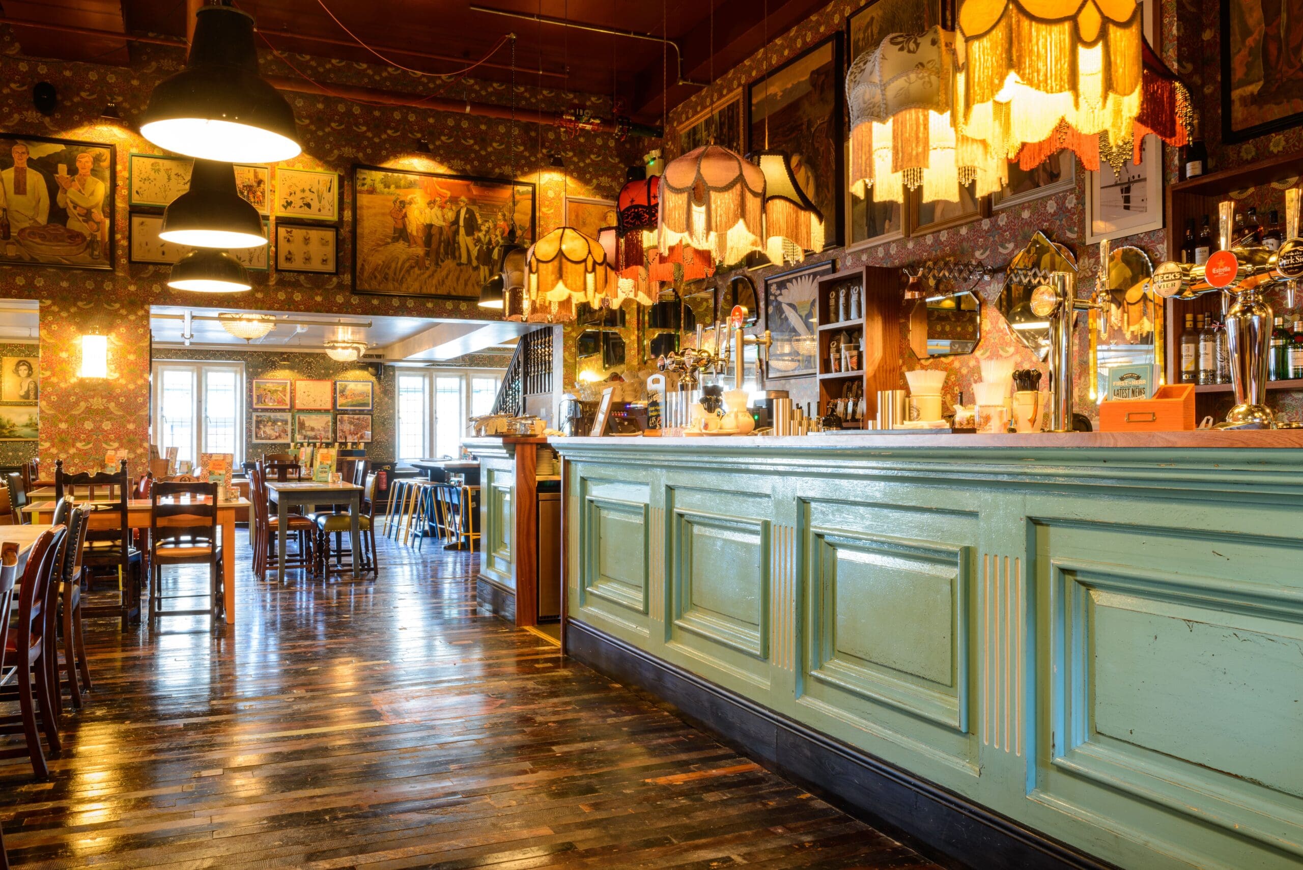 A cozy, vintage-style bar with a turquoise wooden counter, colorful fringed lamps, and wooden floors. Zapato-themed artwork and posters decorate the walls as tables and chairs fill the warmly lit, eclectic space.