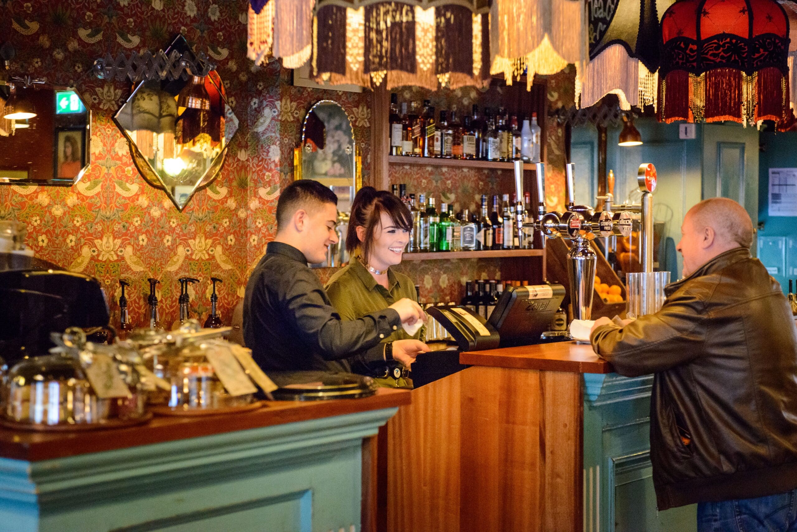 Two bartenders stand behind a bar, smiling and talking with a customer who is leaning on the counter. The vintage Zapato decor features patterned wallpaper, mirrors, and bottles lined up on shelves.
