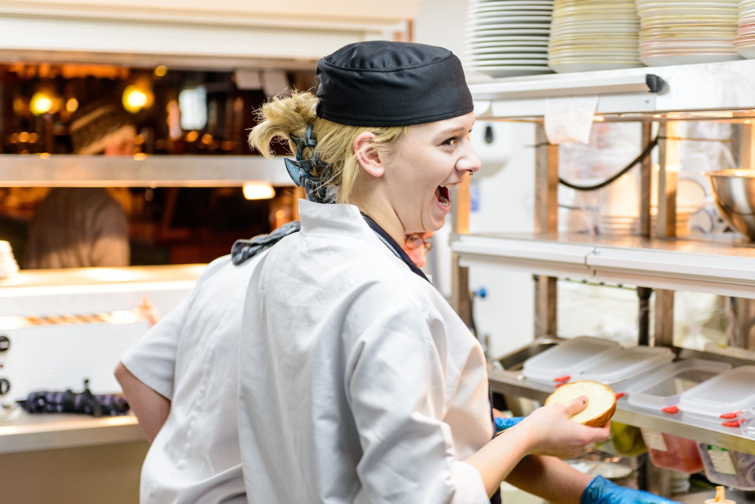 A chef wearing a black hat, white coat, and stylish zapatos smiles widely while holding food in a busy restaurant kitchen, with shelves of plates and another person working beside her.