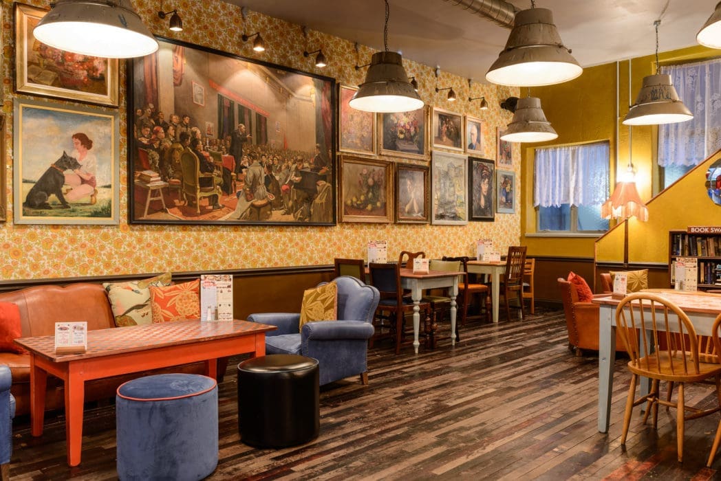 Cozy Argo café interior with mismatched chairs and tables, colorful cushions, wooden floor, floral wallpaper, and numerous framed paintings on the walls. Hanging lamps provide warm lighting, creating a welcoming atmosphere.
