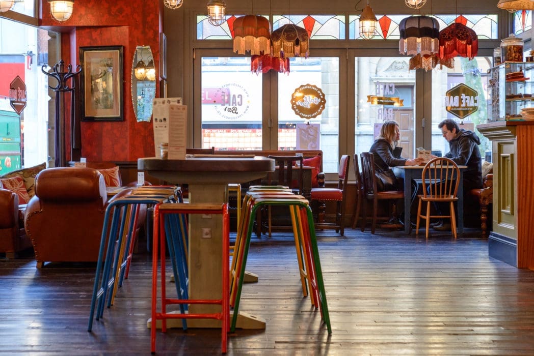 A cozy Argo cafe interior with colorful stools around a tall table, leather couches, and two people sitting by the window having a conversation. Warm lighting and decorative lamps create an inviting atmosphere.