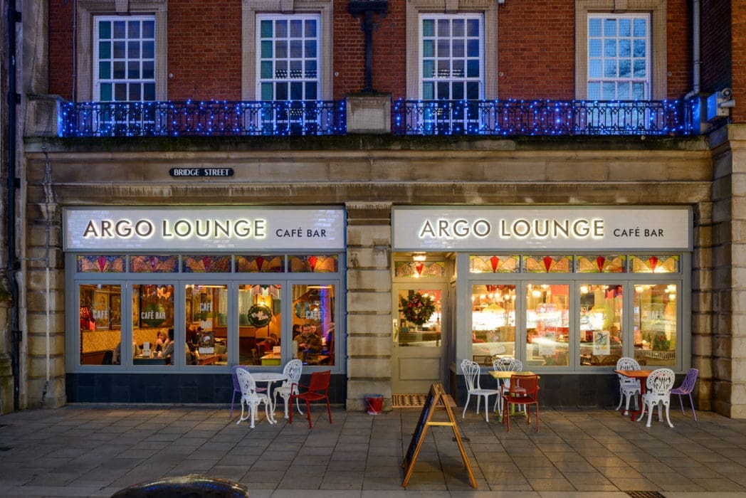 The exterior of Argo shines at night with illuminated signs, large windows revealing a cozy interior, festive holiday decorations, and a few outdoor tables and chairs lining the sidewalk.