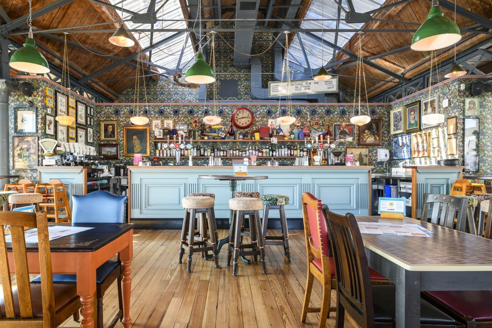 A brightly lit pub interior with wooden floors, patterned walls covered in pictures, and a long Bianco bar with stools. Hanging lights illuminate the mix of wooden tables and chairs set under a high ceiling with skylights.