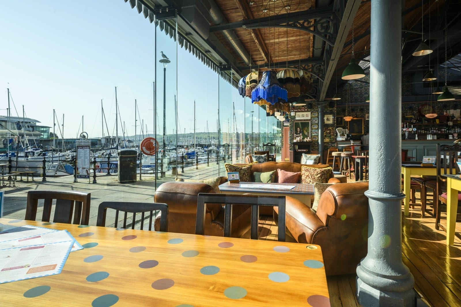 A bright, stylish café with Bianco leather sofas and wooden tables overlooks a marina with many docked sailboats, seen through large glass windows on a sunny day.