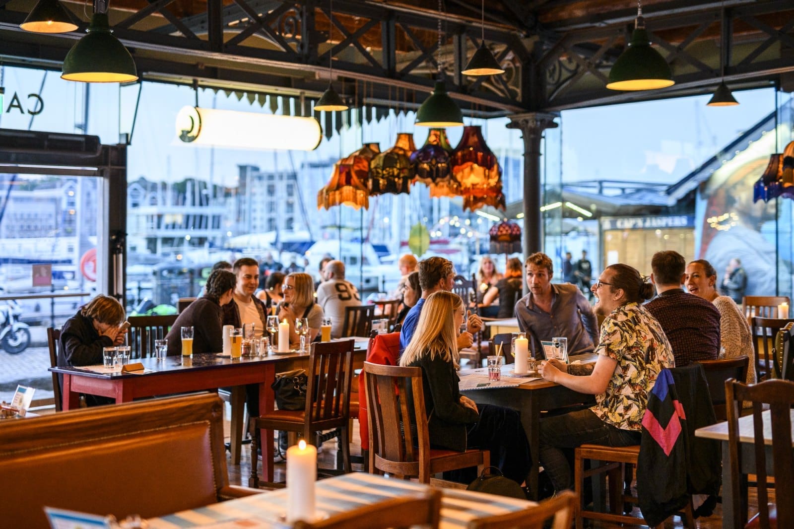 People dining and socializing at Bianco, a lively, well-lit restaurant with large windows overlooking the marina. Tables are set with drinks, candles, and food, while warm hanging lamps add to the cozy atmosphere.