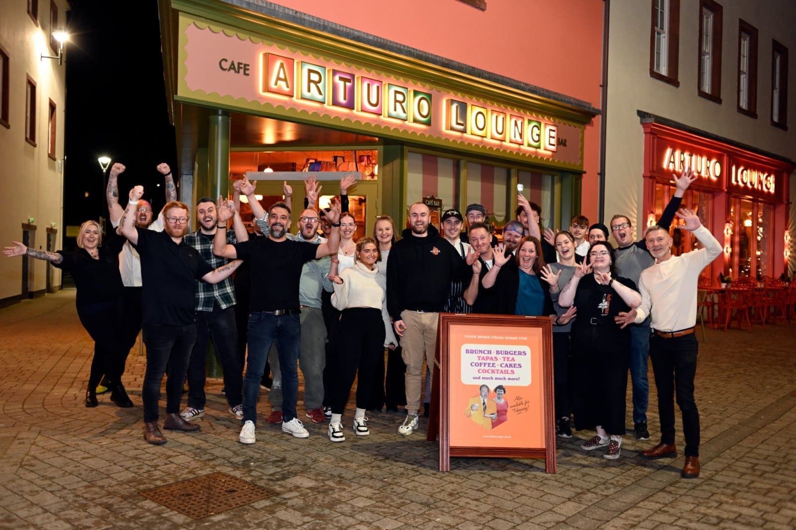 A large group of people stand smiling and waving in front of the brightly-lit Arturo Lounge café at night, with a welcoming Arturo signboard and colorful storefront visible behind them.