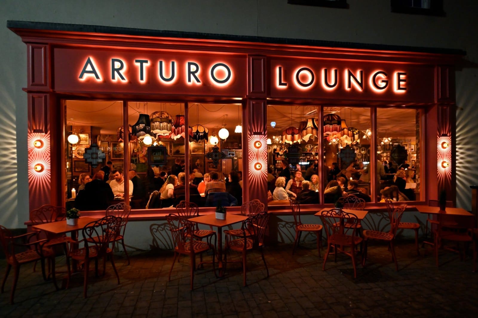 A warmly lit café called Arturo Lounge with red chairs and tables outside, seen at night. Large windows reveal many people inside, sitting and talking under vintage-style hanging lamps in the cozy Arturo atmosphere.