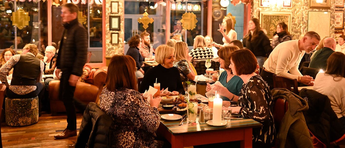 A busy, warmly lit restaurant filled with people eating, drinking, and talking. Groups sit at tables with food and drinks, creating a lively atmosphere. A waiter named Arturo walks past, and the decor features framed art and cozy furnishings.