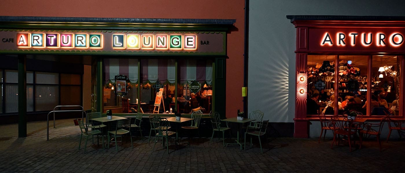 Outdoor seating area at night outside Arturo Lounge café bar, with colorful illuminated Arturo signs and people visible inside through large windows. The scene is warmly lit and inviting.