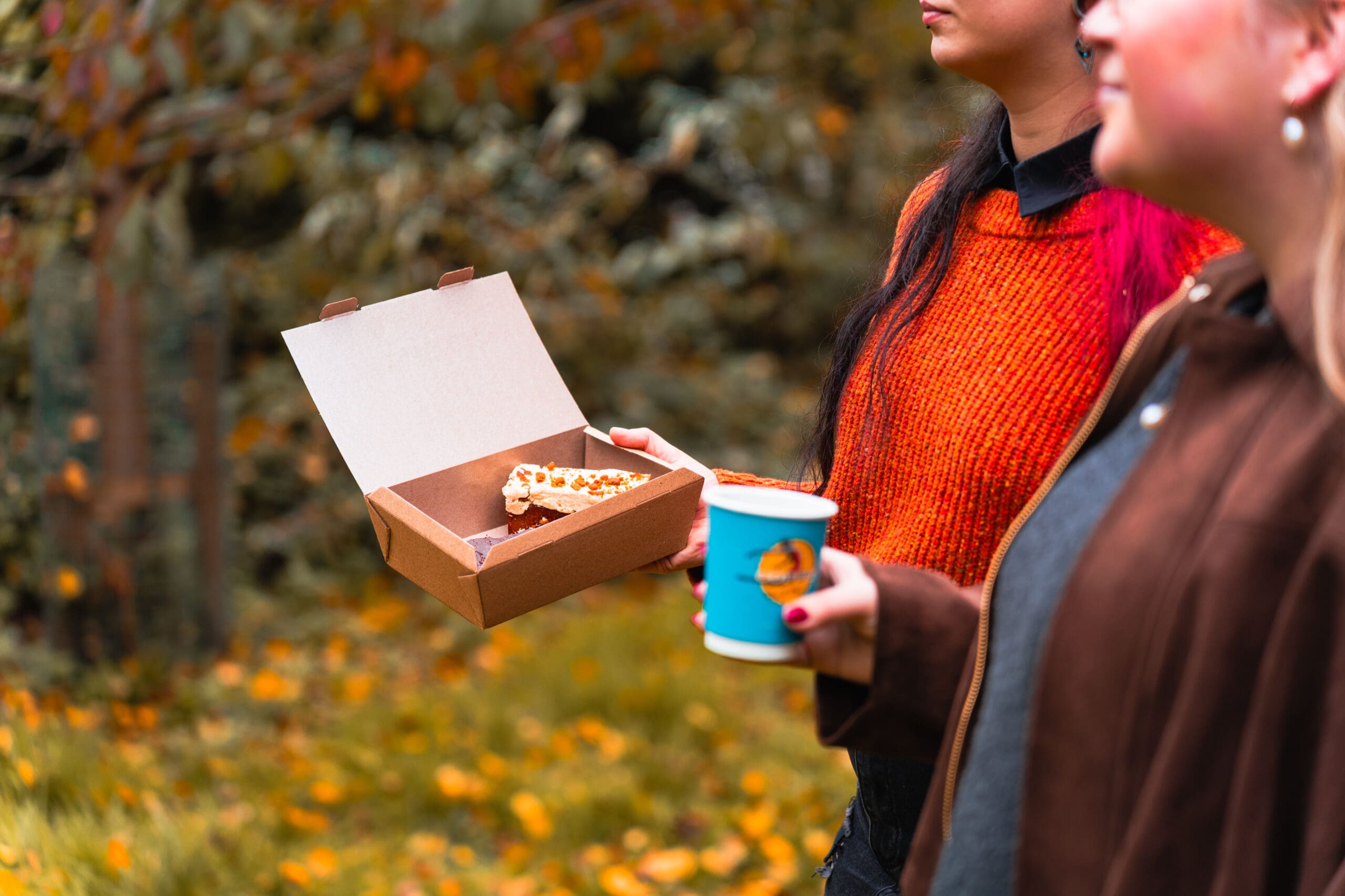 Two people stand outdoors; one holds a paper cup and the other holds an open box with two slices of pizza. Autumn leaves and trees are visible in the background. Their faces are partially out of frame.