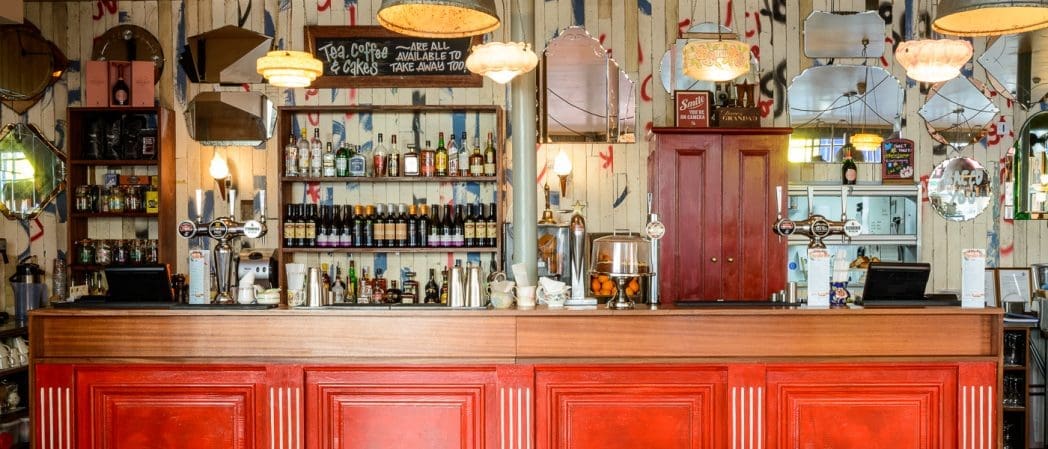 A wooden Bacco bar counter with red panels stands before shelves stocked with bottles, glasses, and drink dispensers. Vintage mirrors and hanging lamps decorate the wall behind the bar.