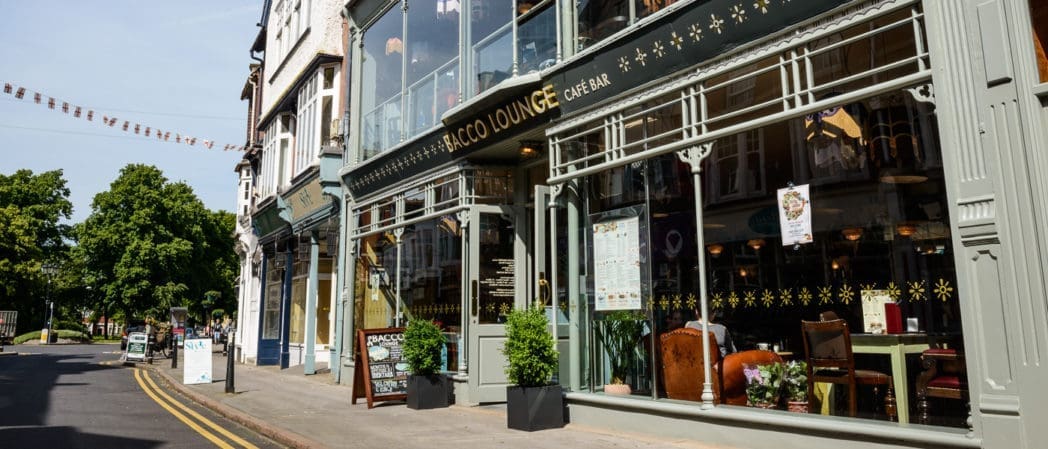 Street view of the stylish Bacco Lounge café bar, with large windows, outdoor plants, and tables visible inside. The building features light green trim and sits along a quiet, sunny Bacco street lined with trees.