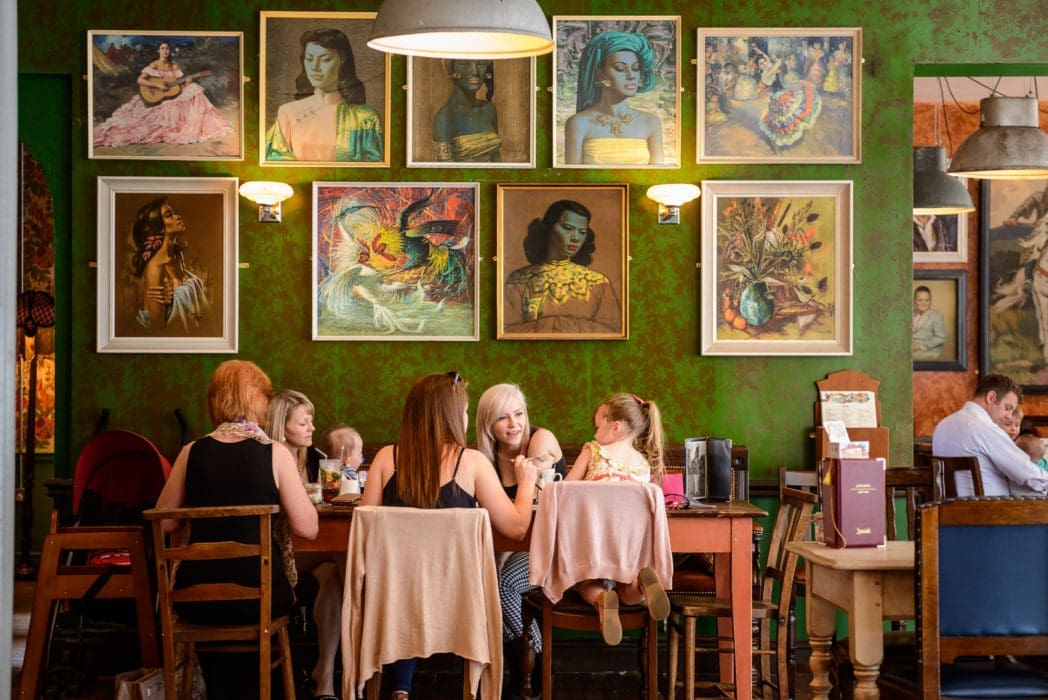 A group of women and children sit around a wooden table in the cozy Bacco cafe, its green textured walls adorned with colorful portraits. Menus and drinks are on the table, with other patrons visible in the background.