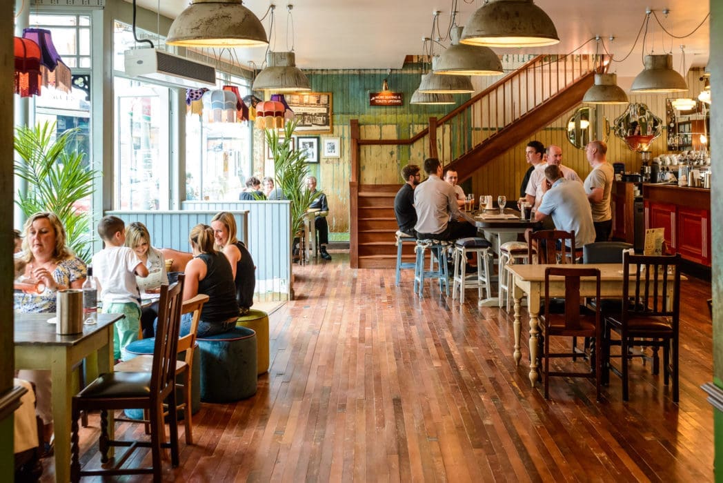 A cozy restaurant with wooden floors, tables, and chairs, Bacco offers a warm, inviting atmosphere. People are dining and chatting near large windows or the bar, while warm lighting and lush plants complete the scene.