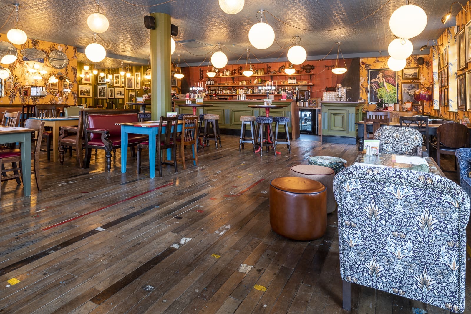 A cozy, eclectic café with wooden floors, various tables and chairs, patterned armchairs, round hanging lights, and a Barco at the back. Walls are decorated with framed pictures and artwork.